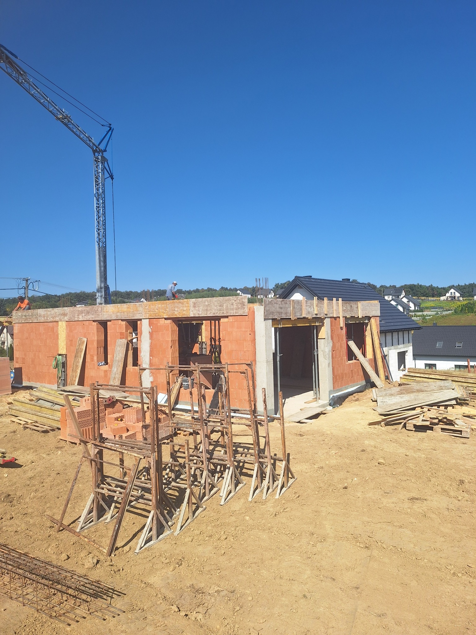 Budowa domu z czerwonej cegły w słoneczny dzień. Widoczny dźwig, rusztowania i pracownicy na dachu. W tle zabudowa jednorodzinna i błękitne niebo.