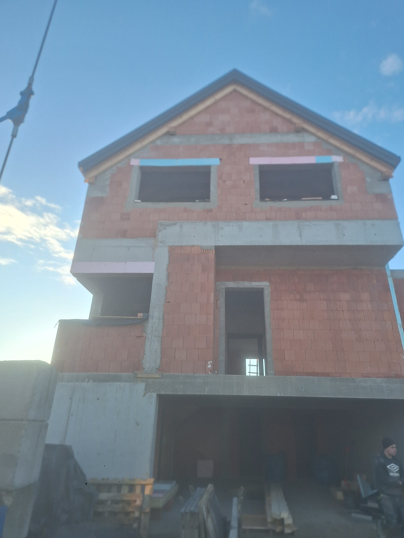 Surowy stan domu murowanego z czerwonej cegły, widoczne betonowe elementy konstrukcyjne, okna zabezpieczone styropianem, garaż w stanie surowym, niebo w tle.