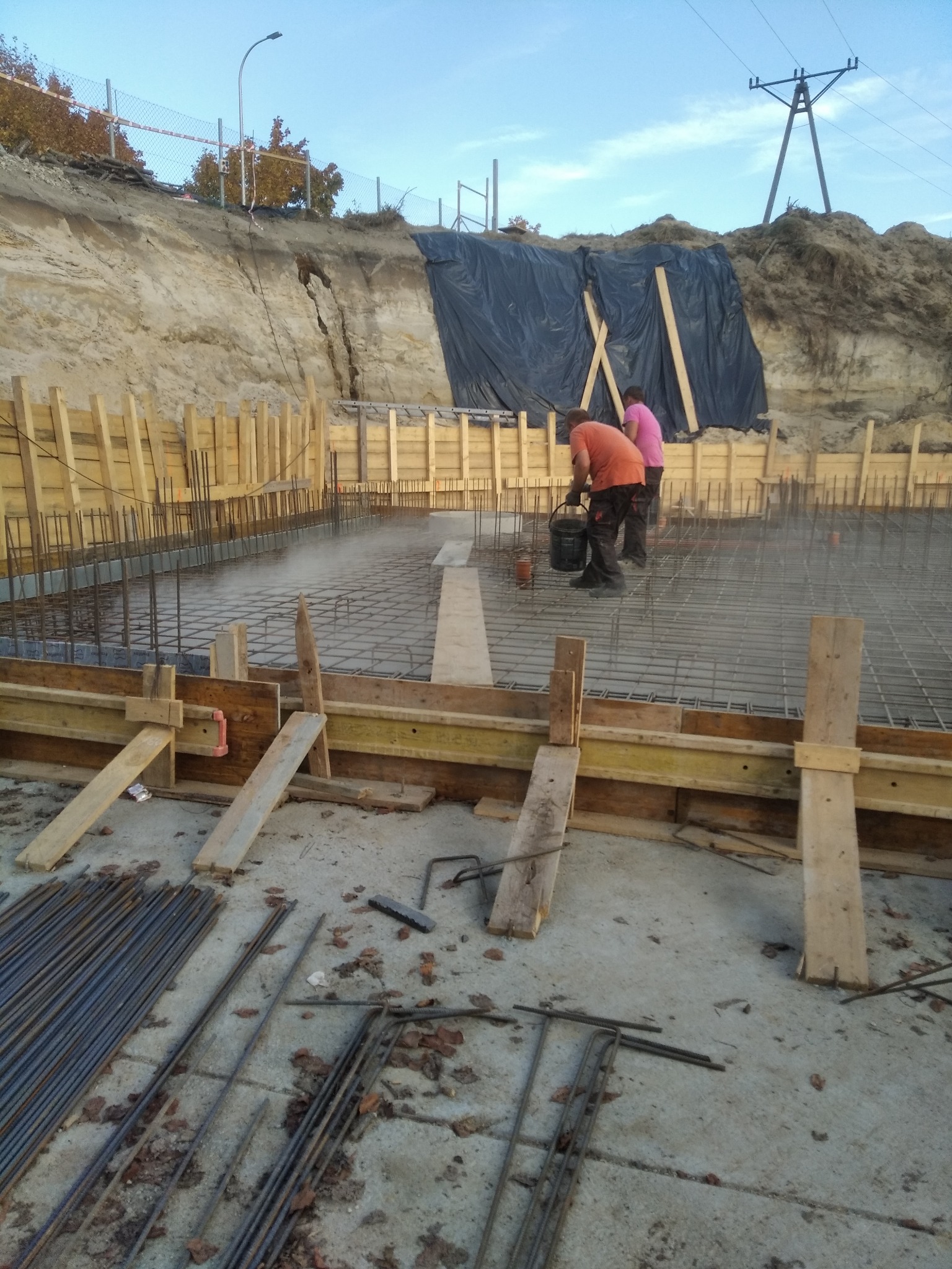 Zbrojenie fundamentów domu z drewnianym szalunkiem i pracownikami w tle, przygotowane do zalania betonem. Widoczne pręty zbrojeniowe i elementy konstrukcyjne.