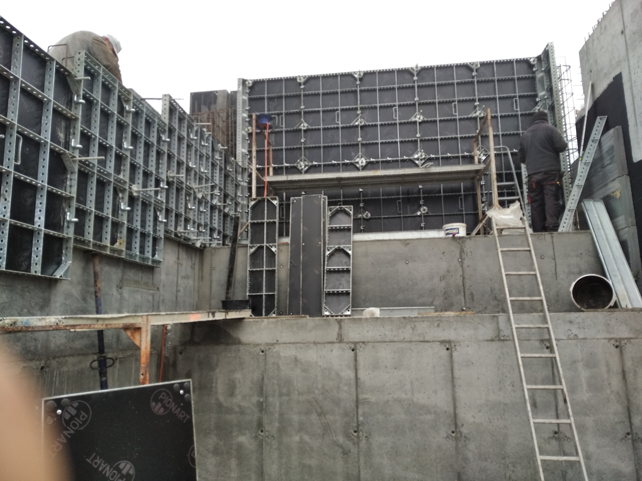 Montaż szalunków Pionart na betonowym fundamencie domu. Widoczni pracownicy i drabina. Szalunki w trakcie instalacji, przygotowanie do wylewania betonu.