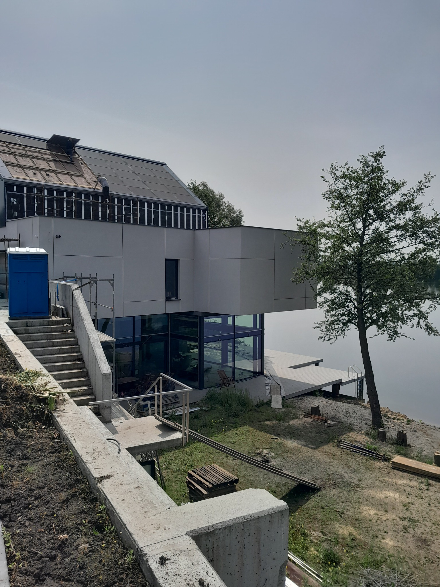 Nowoczesny dom murowany z płaskim dachem w trakcie budowy, widok z zewnątrz na częściowo wykończoną elewację, schody i pomost nad wodą. Na dachu widoczni robotnicy.