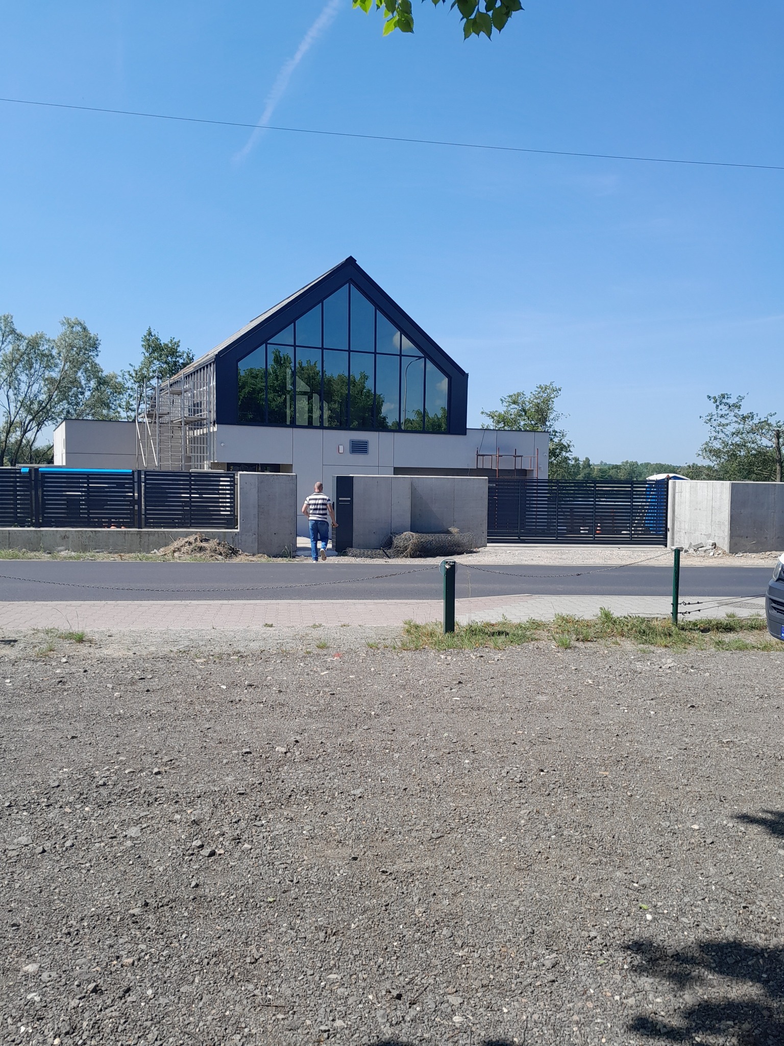 Nowoczesny dom murowany z dużym przeszkleniem szczytowym i czarną stolarką, widoczny od frontu, z osobą przechodzącą obok ogrodzenia. Częściowo widoczne rusztowania.