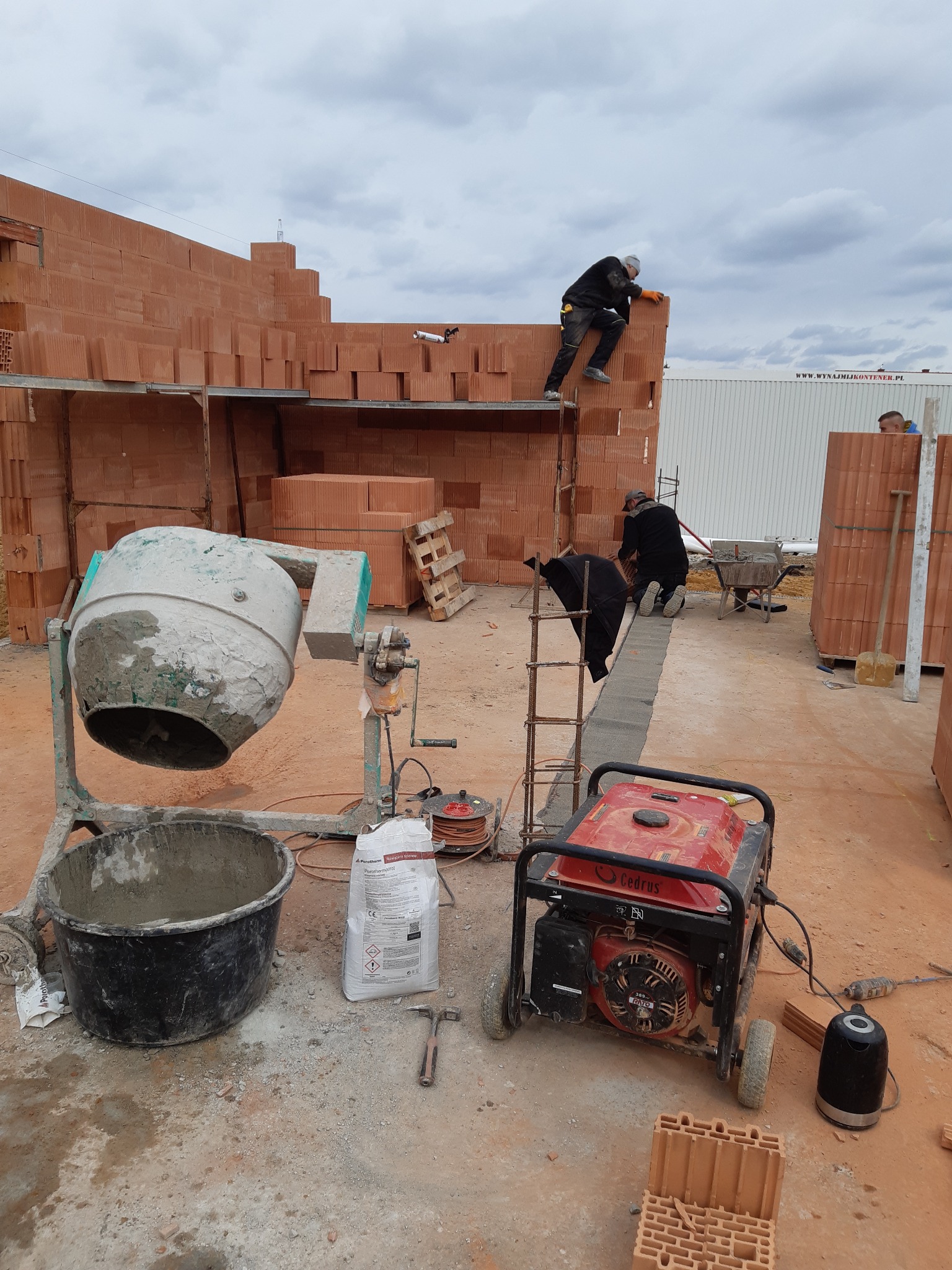 Budowa domu z czerwonej cegły: murarze pracują na rusztowaniu, widoczna betoniarka, generator prądu i materiały budowlane. Etap wznoszenia ścian.