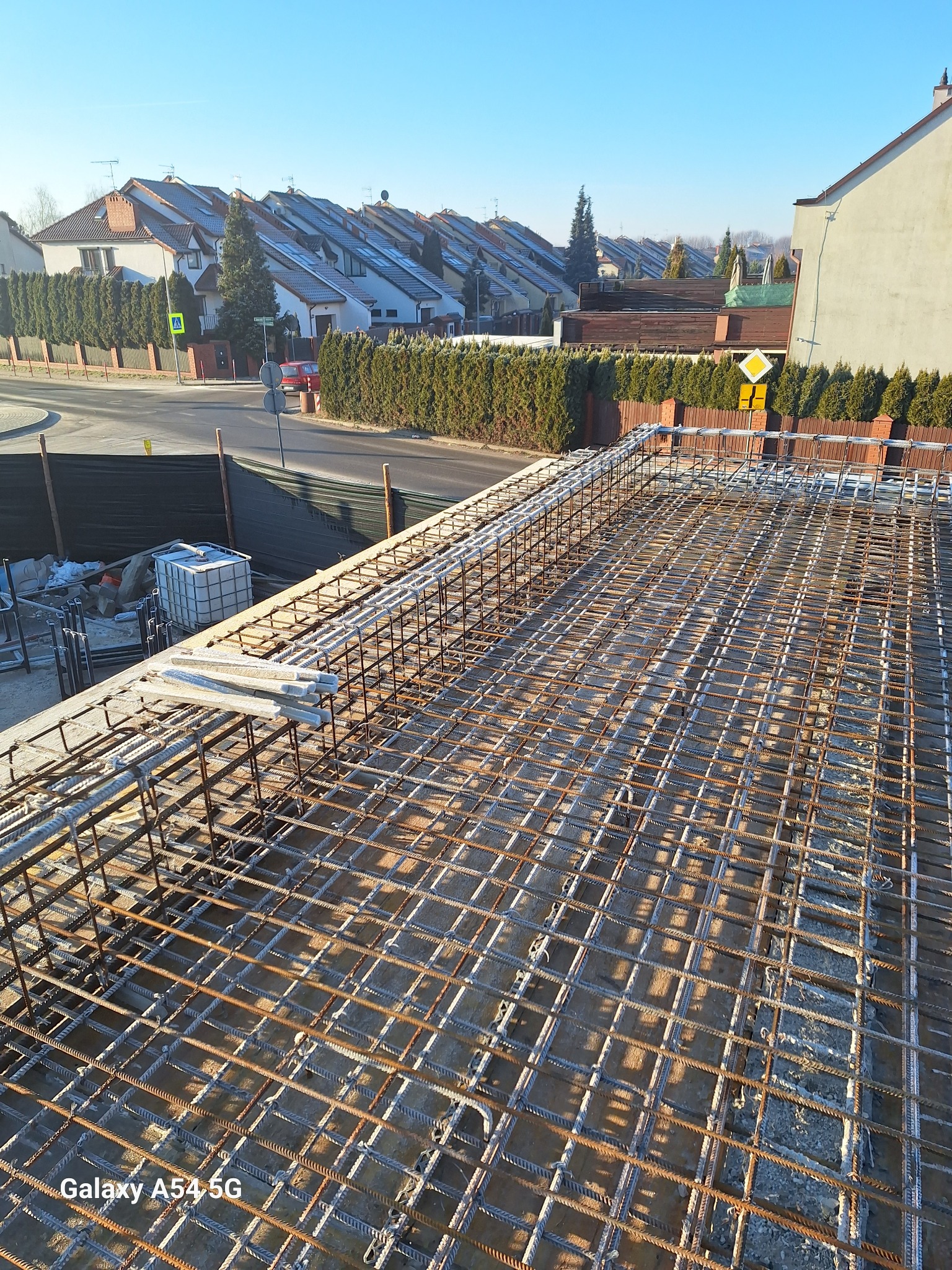 Zbrojenie stropu domu jednorodzinnego w trakcie budowy, widok z góry na siatkę z prętów stalowych, w tle zabudowa osiedlowa i błękitne niebo.