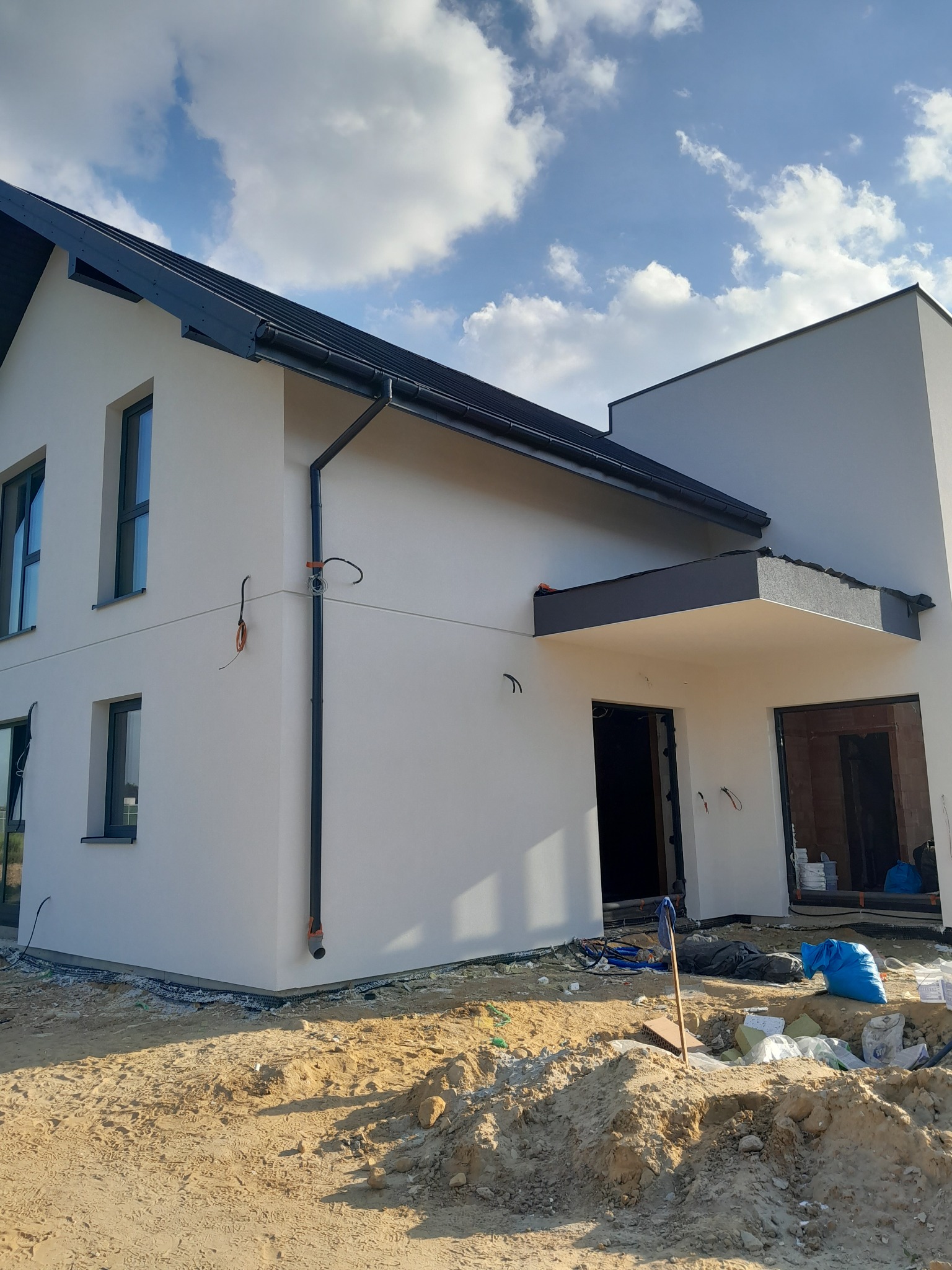 Nowoczesny dom murowany z szarą stolarką okienną i dachem, w trakcie wykończenia elewacji. Widoczne przewody elektryczne i teren budowy.