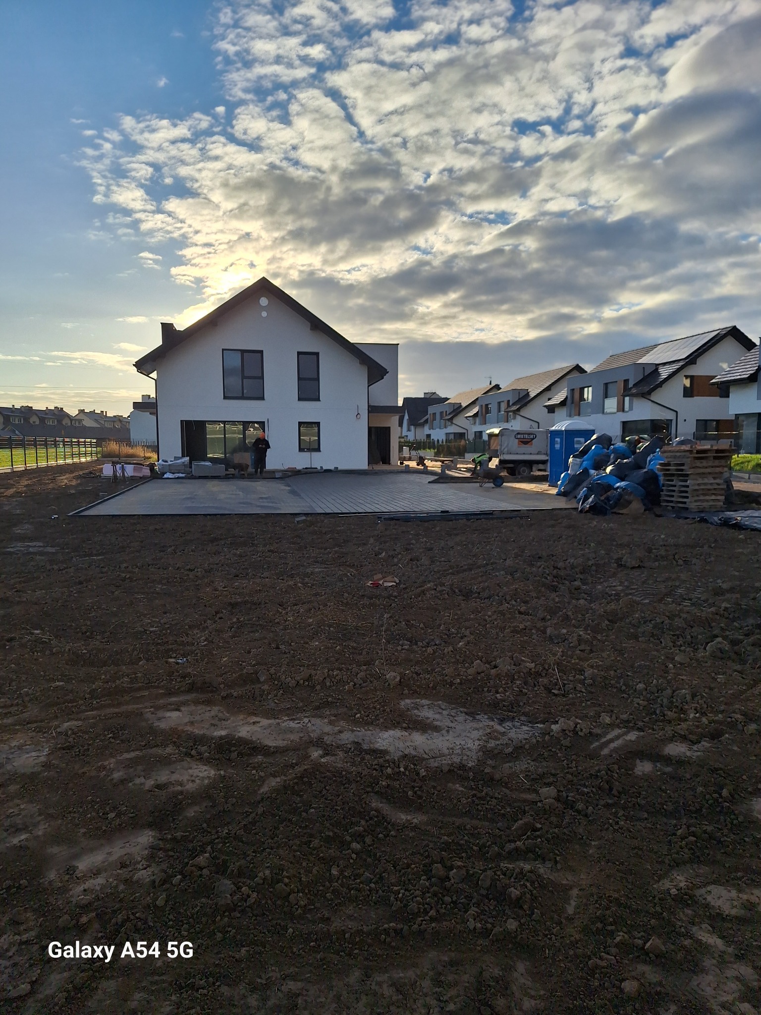 Nowoczesny dom murowany z płaskim dachem w trakcie budowy, widoczne przygotowane podłoże pod taras i pracownicy. W tle błękitne niebo z chmurami.