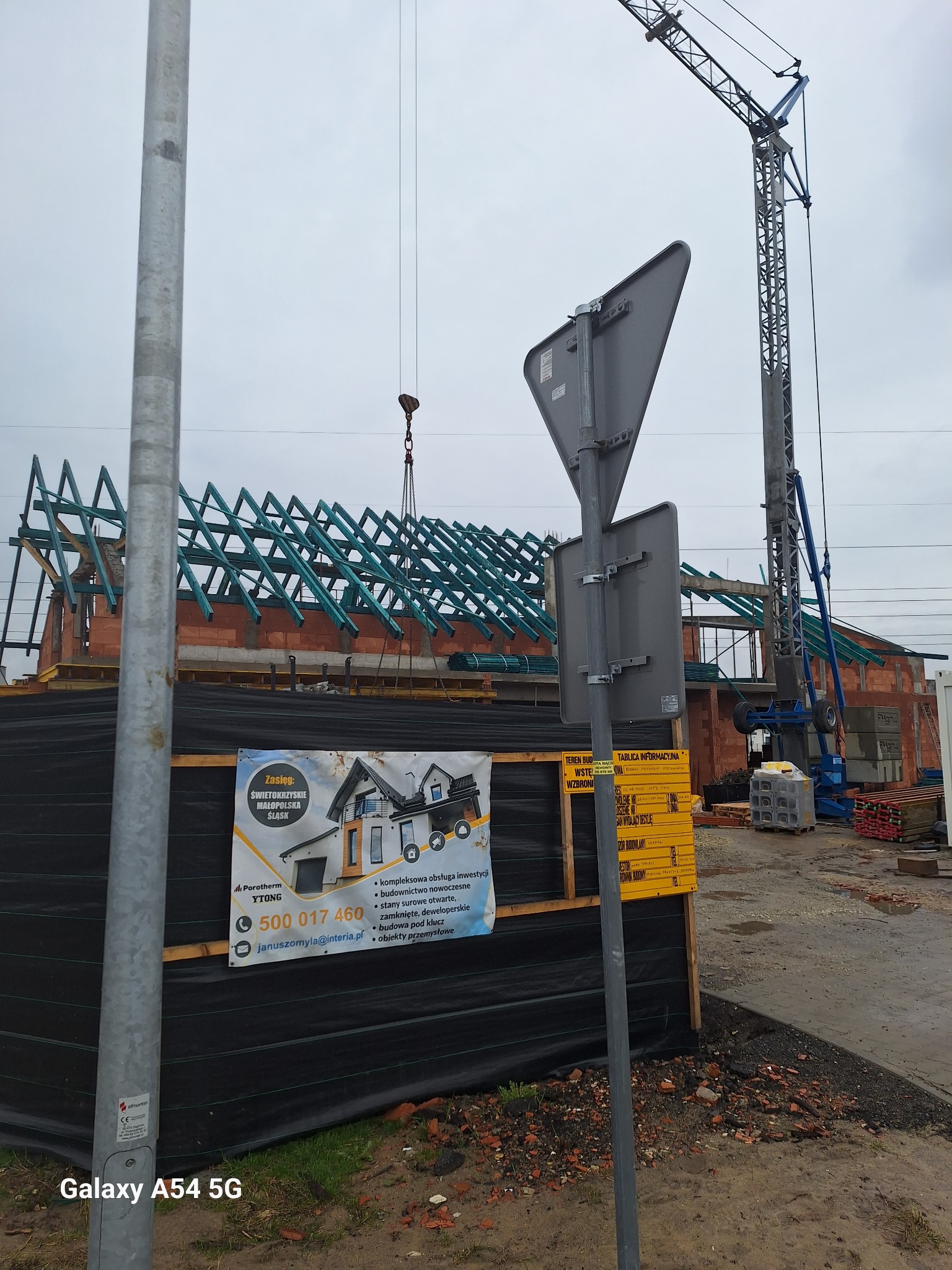 Budowa domu murowanego w trakcie realizacji: ściany z czerwonej cegły, drewniana konstrukcja dachu, żuraw budowlany. Na pierwszym planie tablica informacyjna.