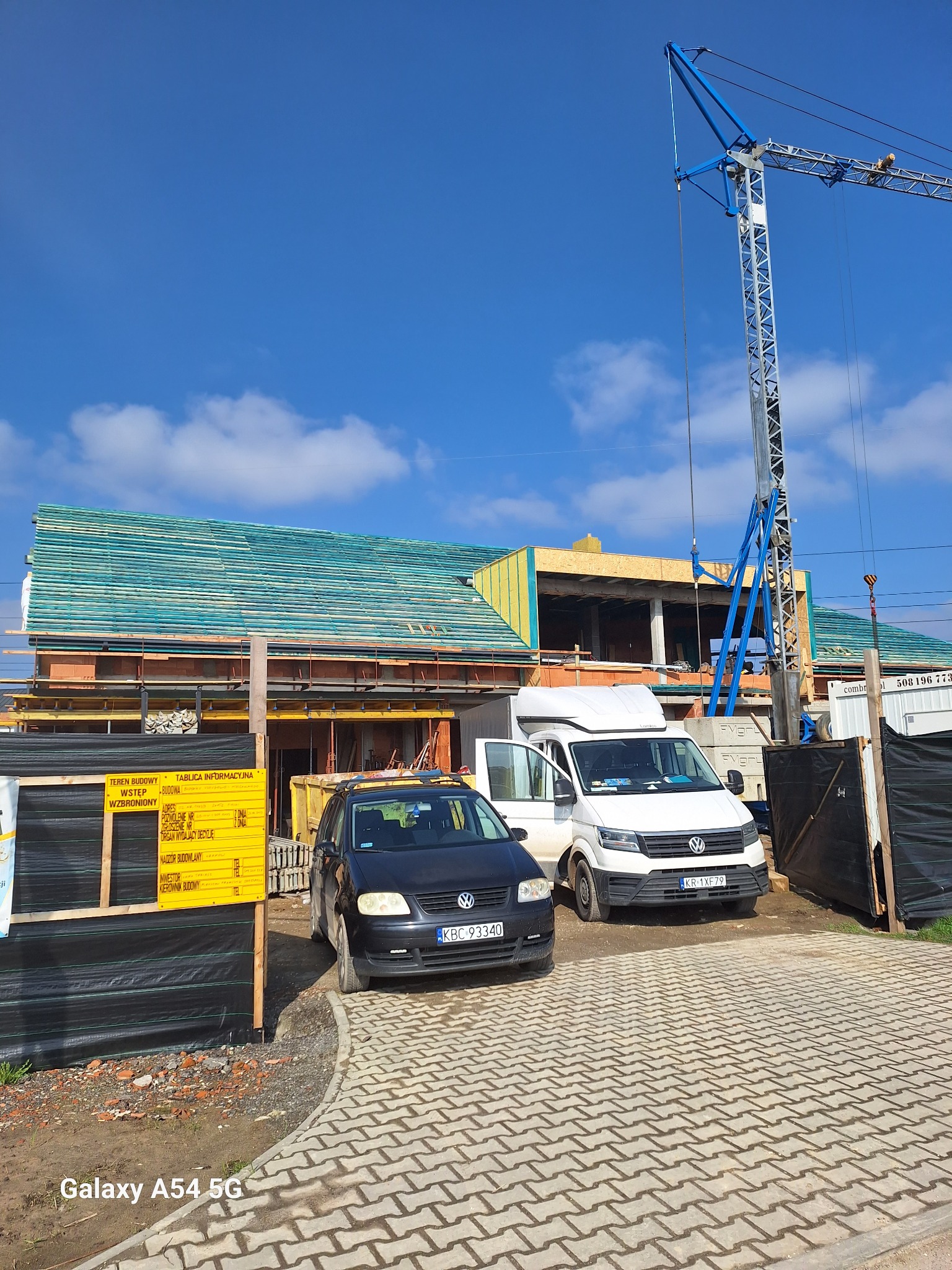 Budowa domu murowanego w trakcie realizacji: widoczny szkielet budynku z cegły, drewniana konstrukcja dachu pokryta folią, dźwig budowlany i zaparkowane samochody.