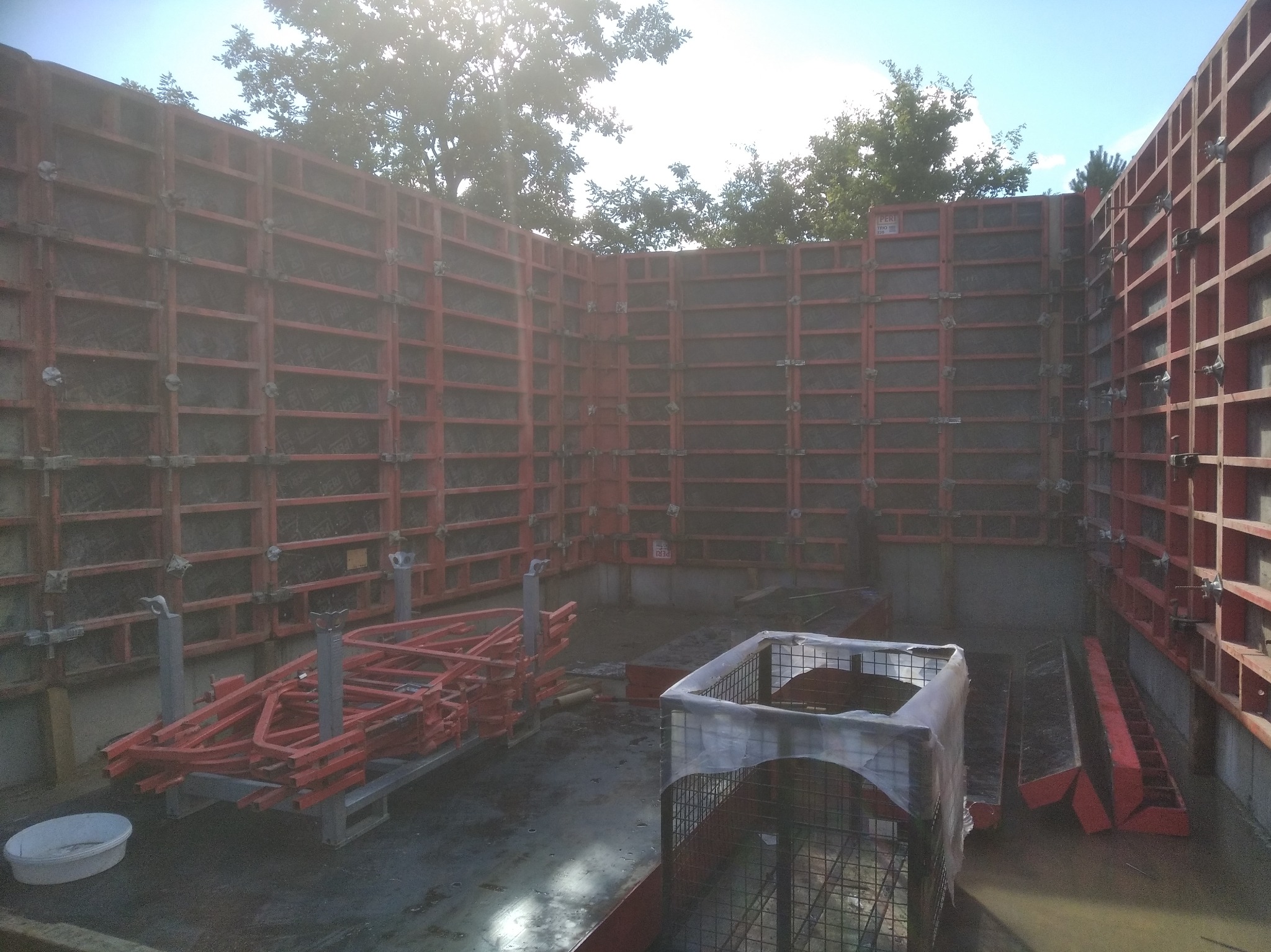 Szare ściany fundamentowe w czerwonym szalunku, przygotowane do zalania betonem. Widoczne elementy konstrukcyjne i akcesoria budowlane. Ujęcie z dołu, w tle drzewa.