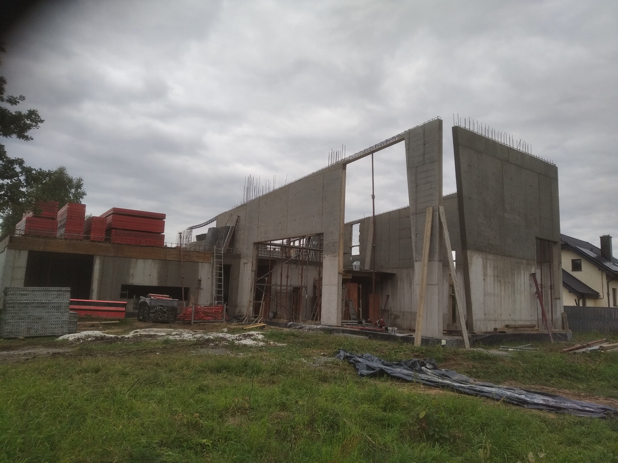 Surowy stan domu murowanego w trakcie budowy: betonowe ściany z wystającymi prętami zbrojeniowymi, szalunki i rusztowania. W tle widoczny fragment gotowego domu.