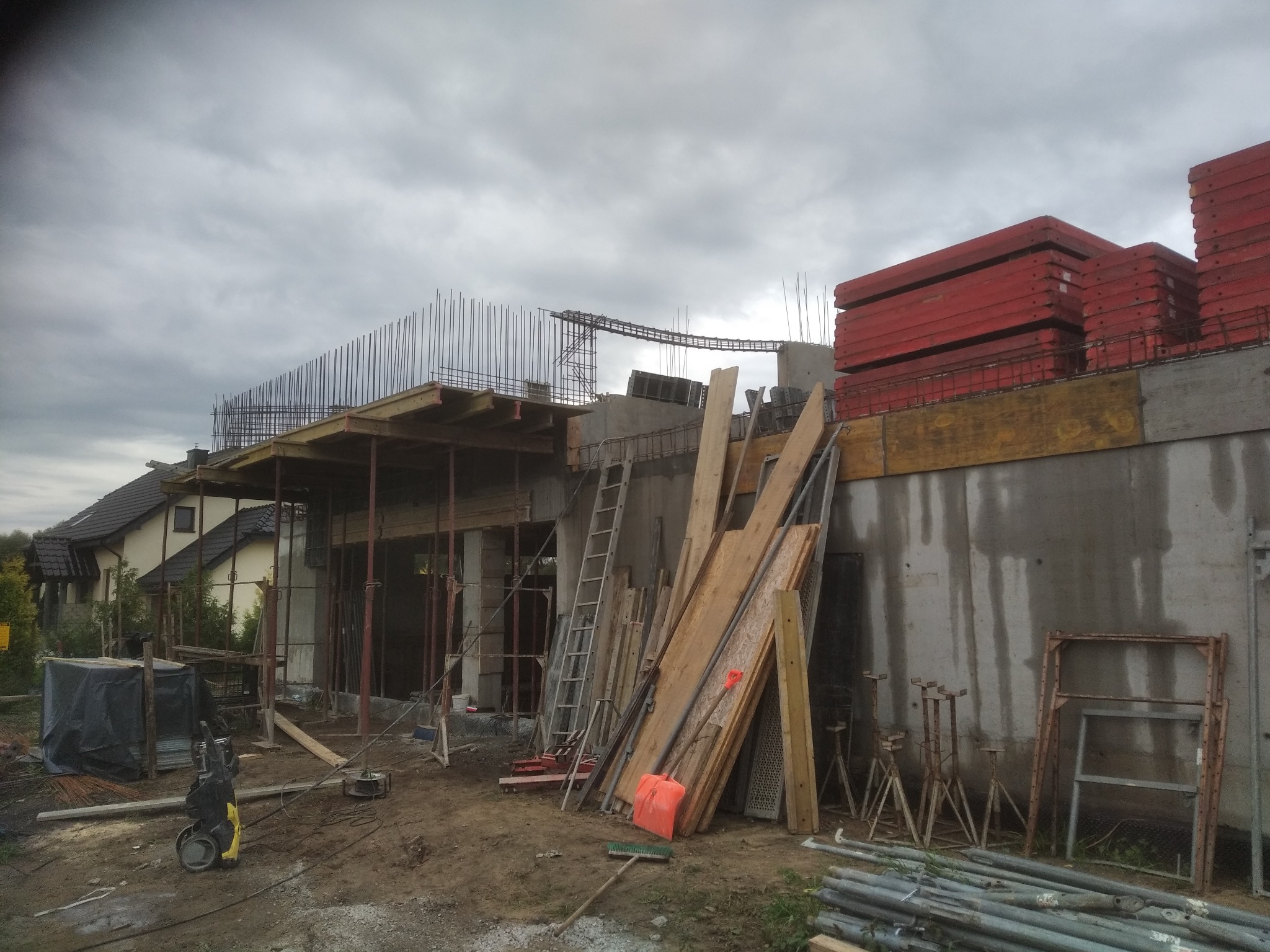 Budowa domu w stanie surowym otwartym: betonowe ściany, zbrojenia, drewniane szalunki, drabina i narzędzia budowlane na placu budowy pod pochmurnym niebem.