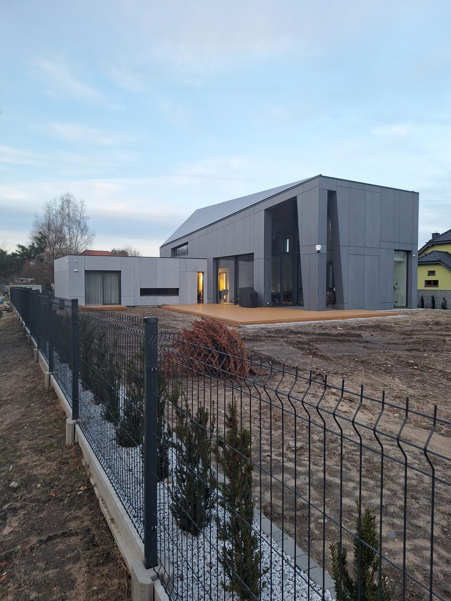 Nowoczesny dom murowany z szarą elewacją i drewnianym tarasem, otoczony metalowym ogrodzeniem z nasadzeniami. Widok z zewnątrz, w tle błękitne niebo.