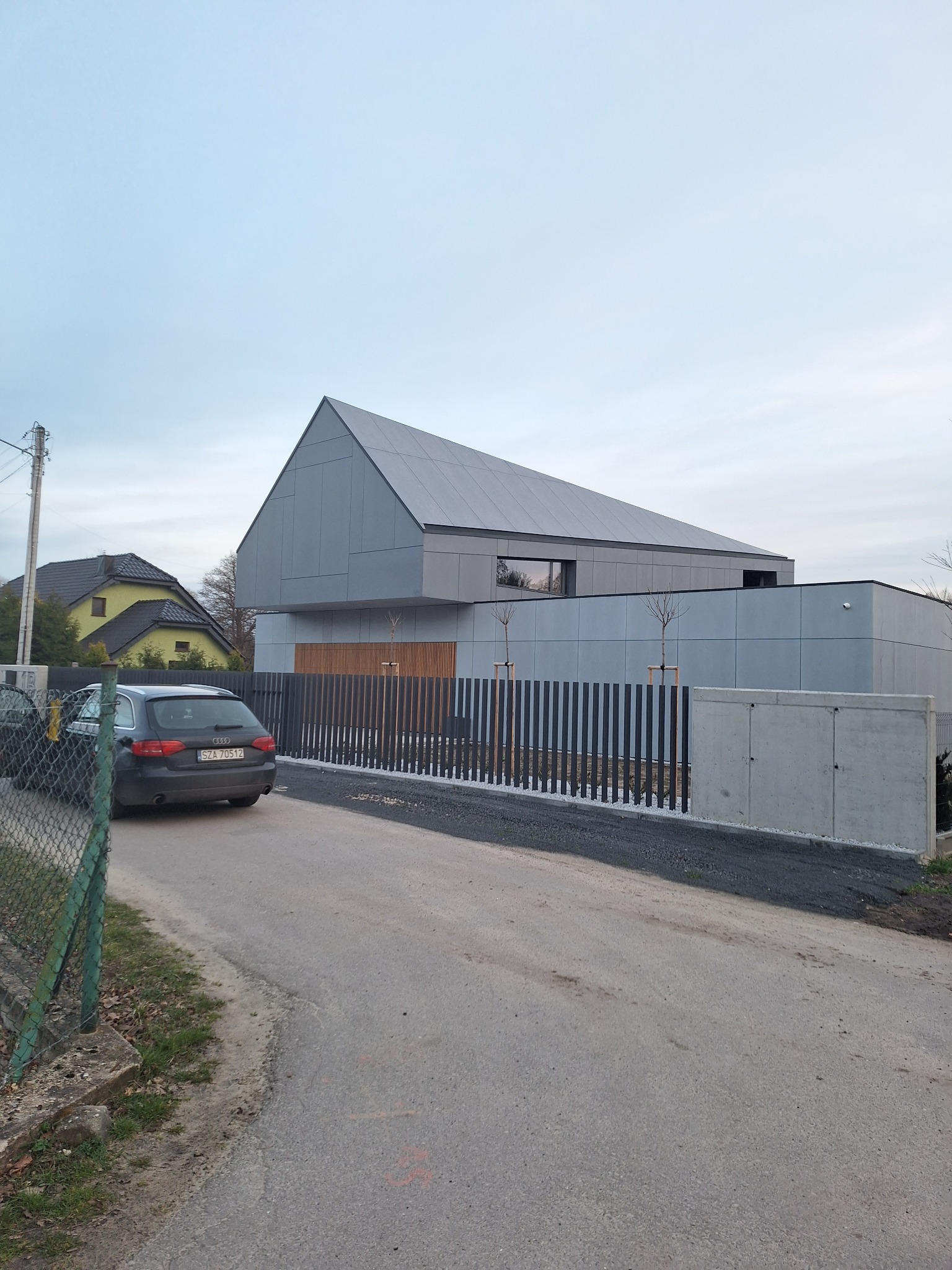 Nowoczesny dom murowany z szarą elewacją i dachem w kształcie trapezu, ogrodzony metalowym płotem. Na podjeździe stoi ciemny samochód osobowy.