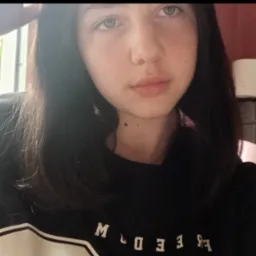 Portret młodej kobiety z ciemnymi włosami, w czarnej bluzie z białym napisem, na tle różowej ściany i okna z zielenią. Ujęcie z góry, z delikatnym oświetleniem.