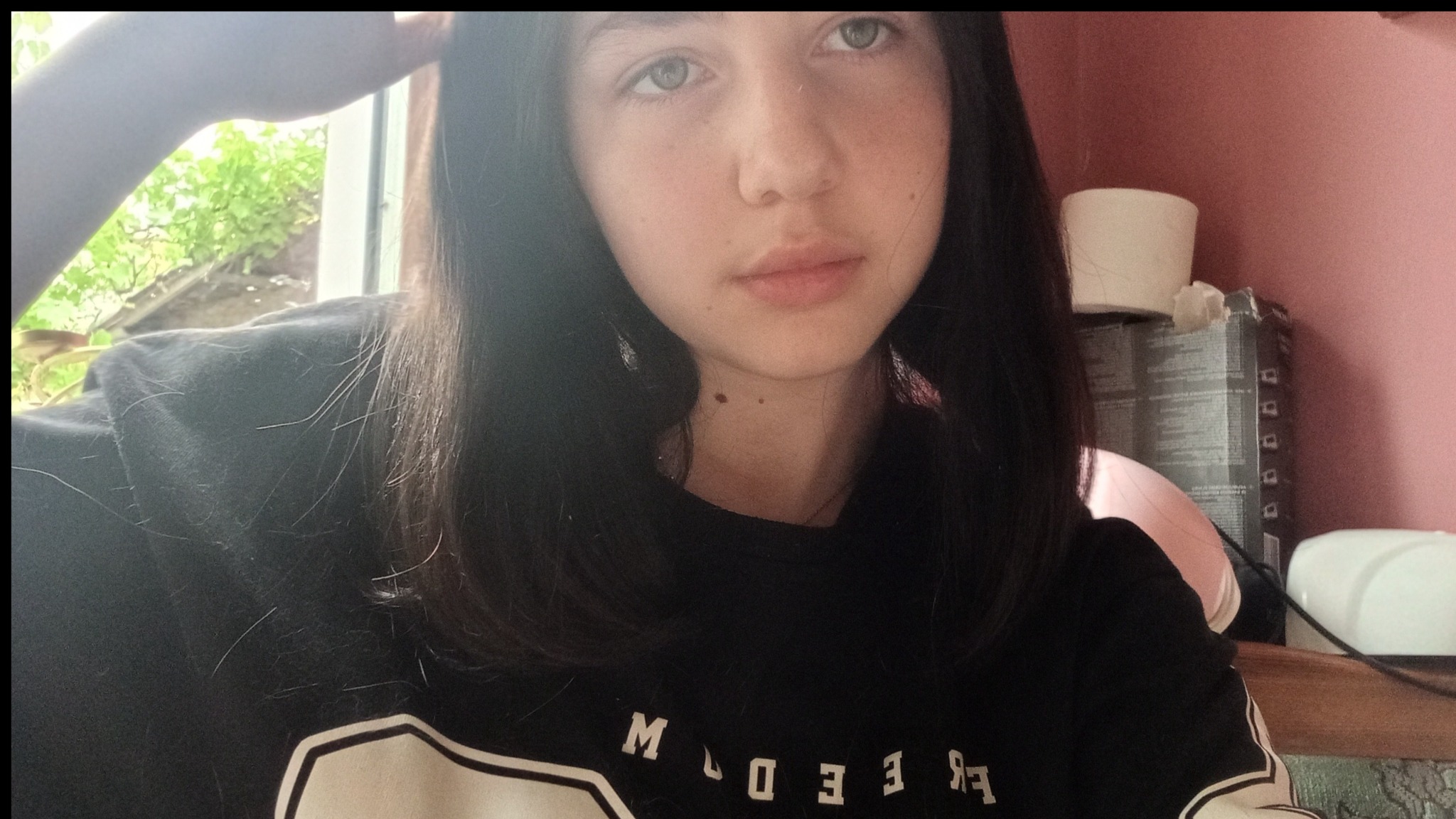 Portret młodej kobiety z ciemnymi włosami, w czarnej bluzie z białym napisem, na tle różowej ściany i okna z zielenią. Ujęcie z góry, z delikatnym oświetleniem.