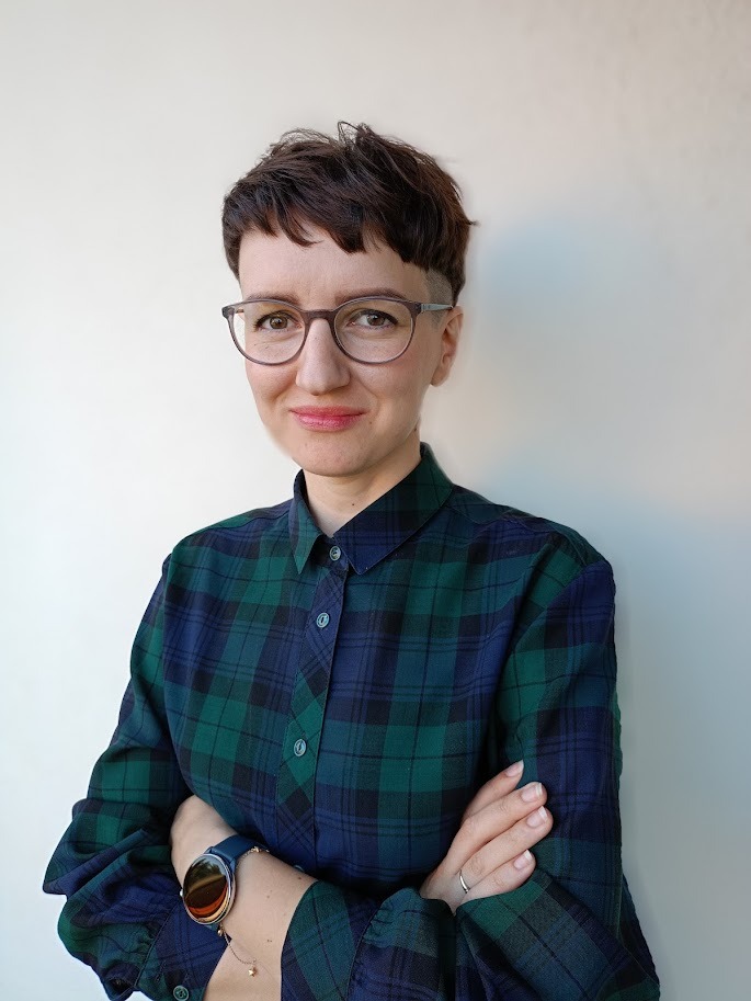 Portret kobiety w okularach i koszuli w kratę, z zegarkiem na ręce, na jasnym tle. Uśmiechnięta, z rękami skrzyżowanymi na piersiach. Krótkie, ciemne włosy.