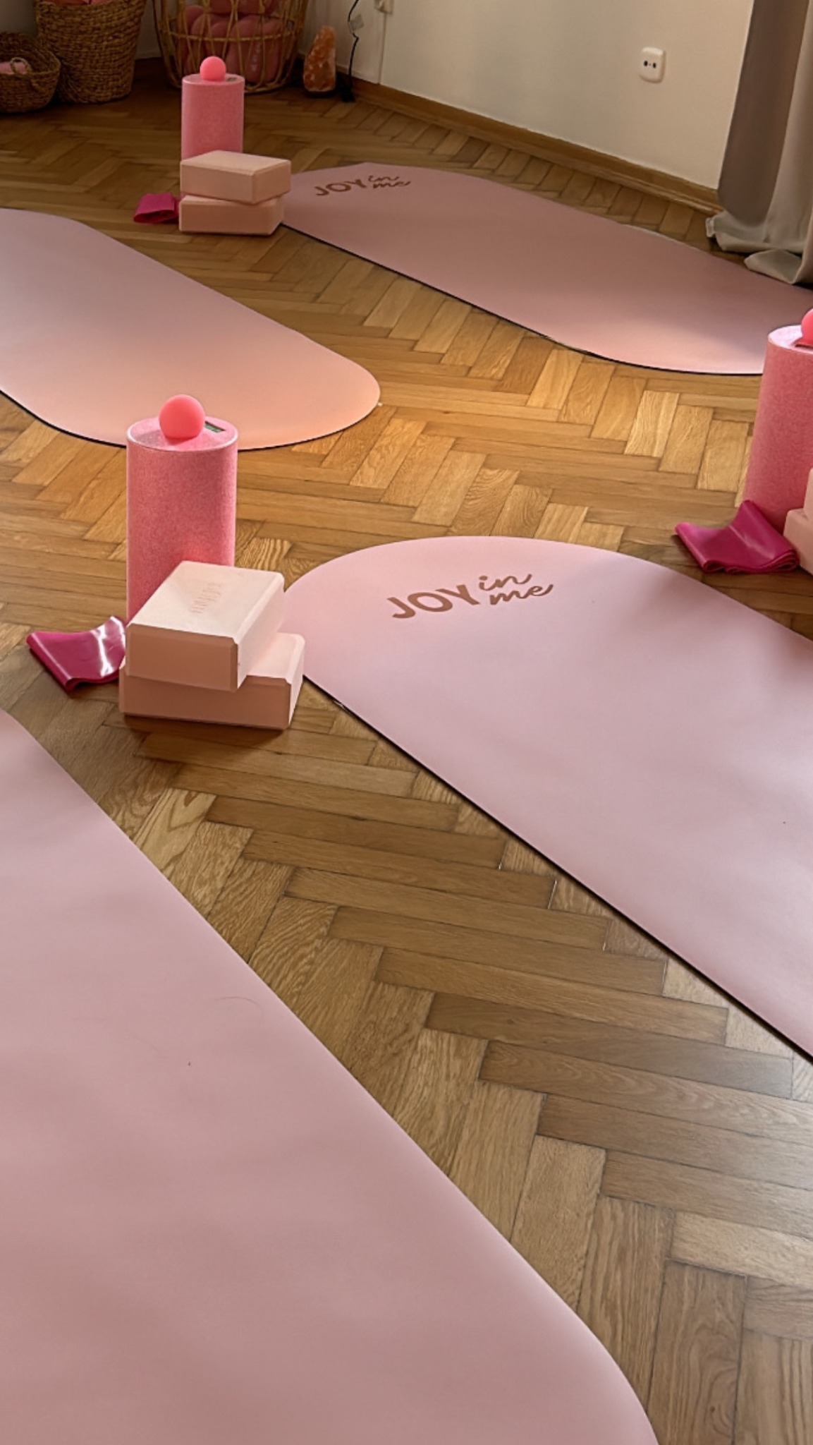 Studio jogi: różowe maty z logo, bloki i wałki na parkiecie w jodełkę. Przytulne wnętrze z elementami dekoracyjnymi i naturalnym światłem.