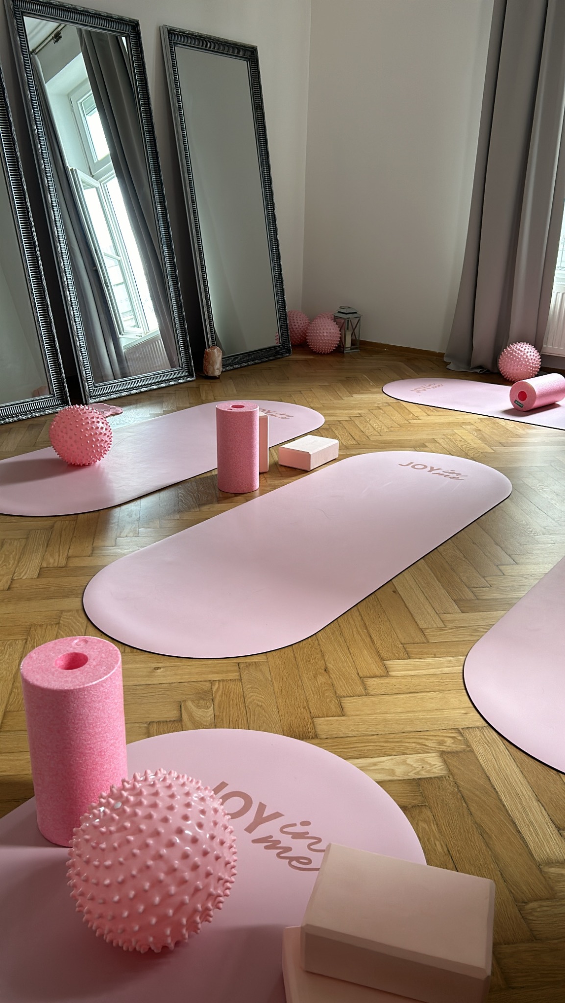 Jasne studio jogi z różowymi matami, wałkami i piłkami do masażu na parkiecie. Dwa lustra w czarnych ramach i szare zasłony w tle.