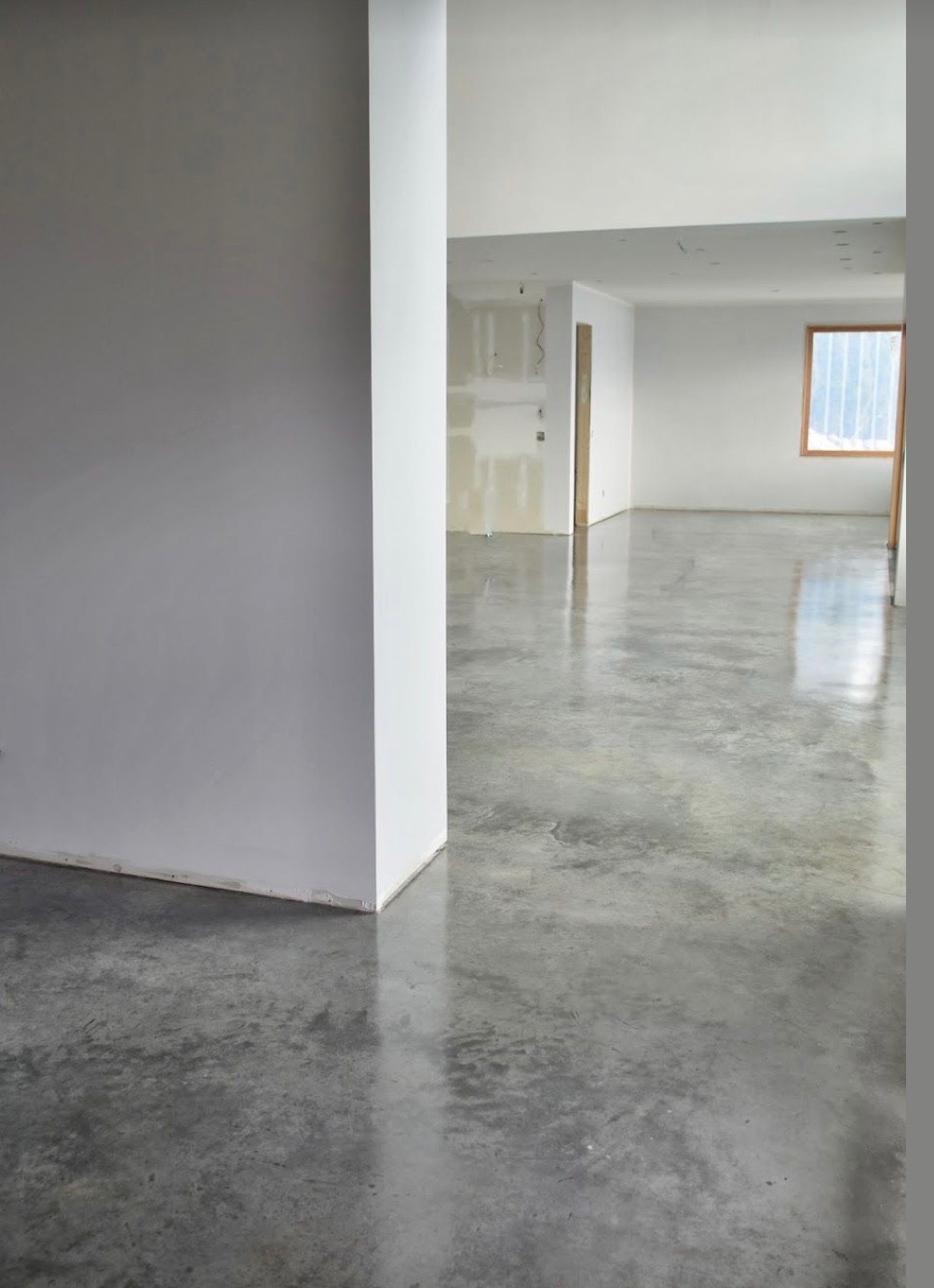 Szara, błyszcząca posadzka w jasnym wnętrzu. Widoczna kolumna i fragmenty ścian. Podłoga odbija światło, tworząc efekt lustrzany. W tle okno i przygotowane ściany do malowania.