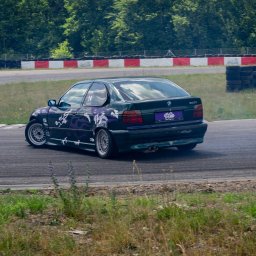 Jakub Sak - Zielony samochód BMW 316i z fioletowymi elementami graficznymi driftuje na torze wyścigowym, z widocznym dymem spod kół i zielonym tłem drzew.