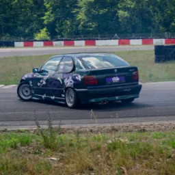 Zielony samochód BMW 316i z fioletowymi elementami graficznymi driftuje na torze wyścigowym, z widocznym dymem spod kół i zielonym tłem drzew.