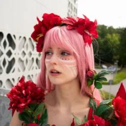 Portret kobiety z różowymi włosami, wiankiem z czerwonych kwiatów i białym makijażem na twarzy, na tle nowoczesnego budynku i zieleni.