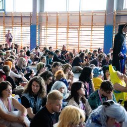 Jakub Sak - Spora grupa osób siedzi na podłodze w hali sportowej, prawdopodobnie podczas wydarzenia cosplay lub konwentu. W tle widoczne okna i drabinki gimnastyczne.