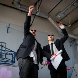 Jakub Sak - Ekscytacja w Białymstoku: Dwóch elegancko ubranych mężczyzn w okularach przeciwsłonecznych celebruje sukces, unosząc ręce w górę na tle sali gimnastycznej.