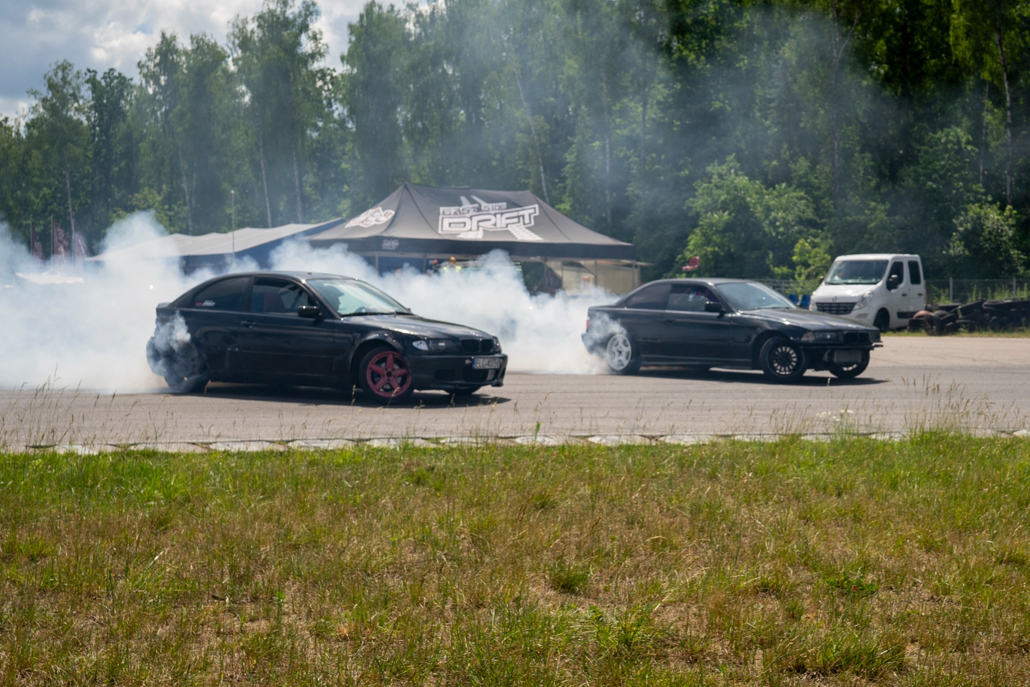 Dwa czarne samochody driftują na torze, unosząc tumany dymu, w tle namiot z napisem 'East Side Drift' i biały van. Dynamiczne ujęcie z zawodów driftingowych.