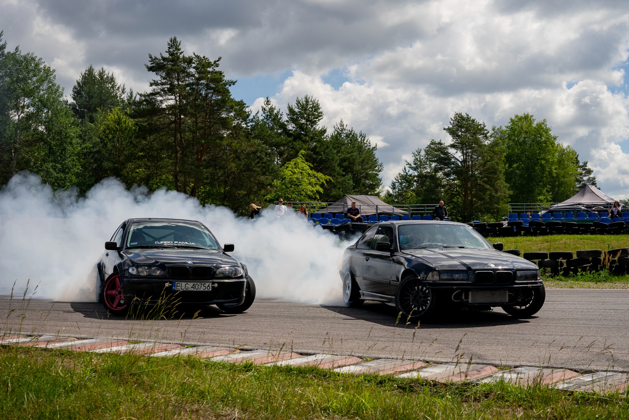 Dwa czarne samochody BMW E36 driftują na torze, unosząc tumany dymu. W tle widoczni widzowie i zieleń drzew.