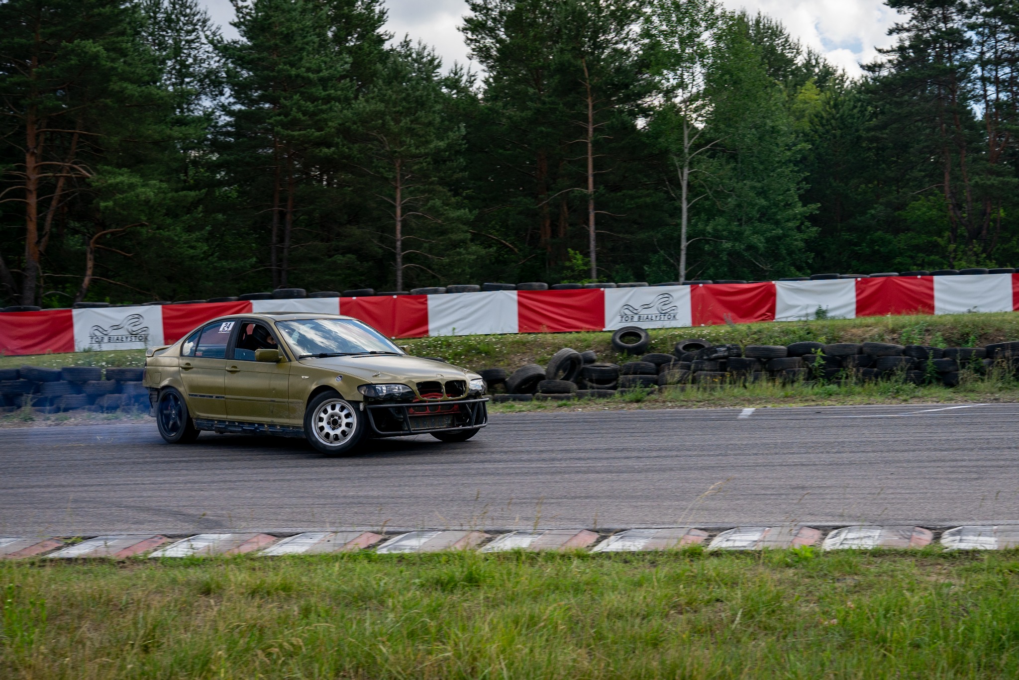 Złoty samochód BMW E46 driftuje na torze wyścigowym, unosząc dym z opon. Widoczny fragment toru z oponami i banerami reklamowymi.