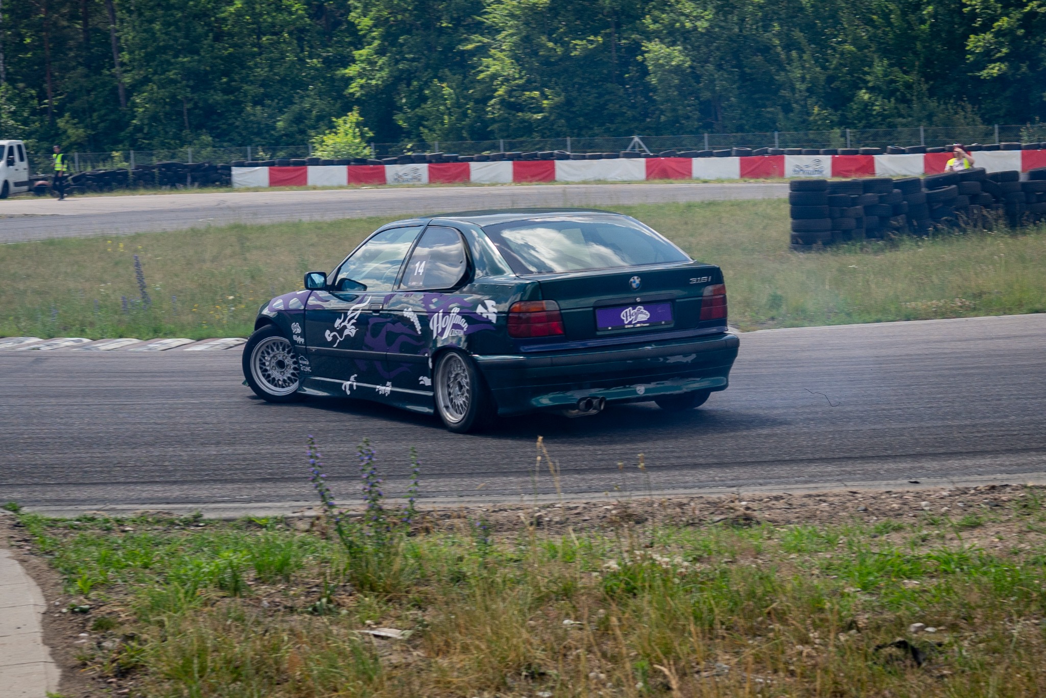 Zielony samochód BMW 316i z fioletowymi elementami graficznymi driftuje na torze wyścigowym, z widocznym dymem spod kół i zielonym tłem drzew.