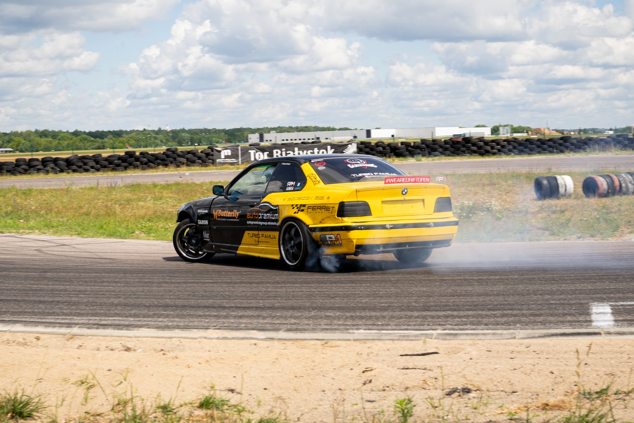 Żółto-czarne BMW E36 driftuje na torze, z opon unosi się dym. Widoczne logotypy sponsorów i napis 'Tor Białystok'. W tle opony i budynki.