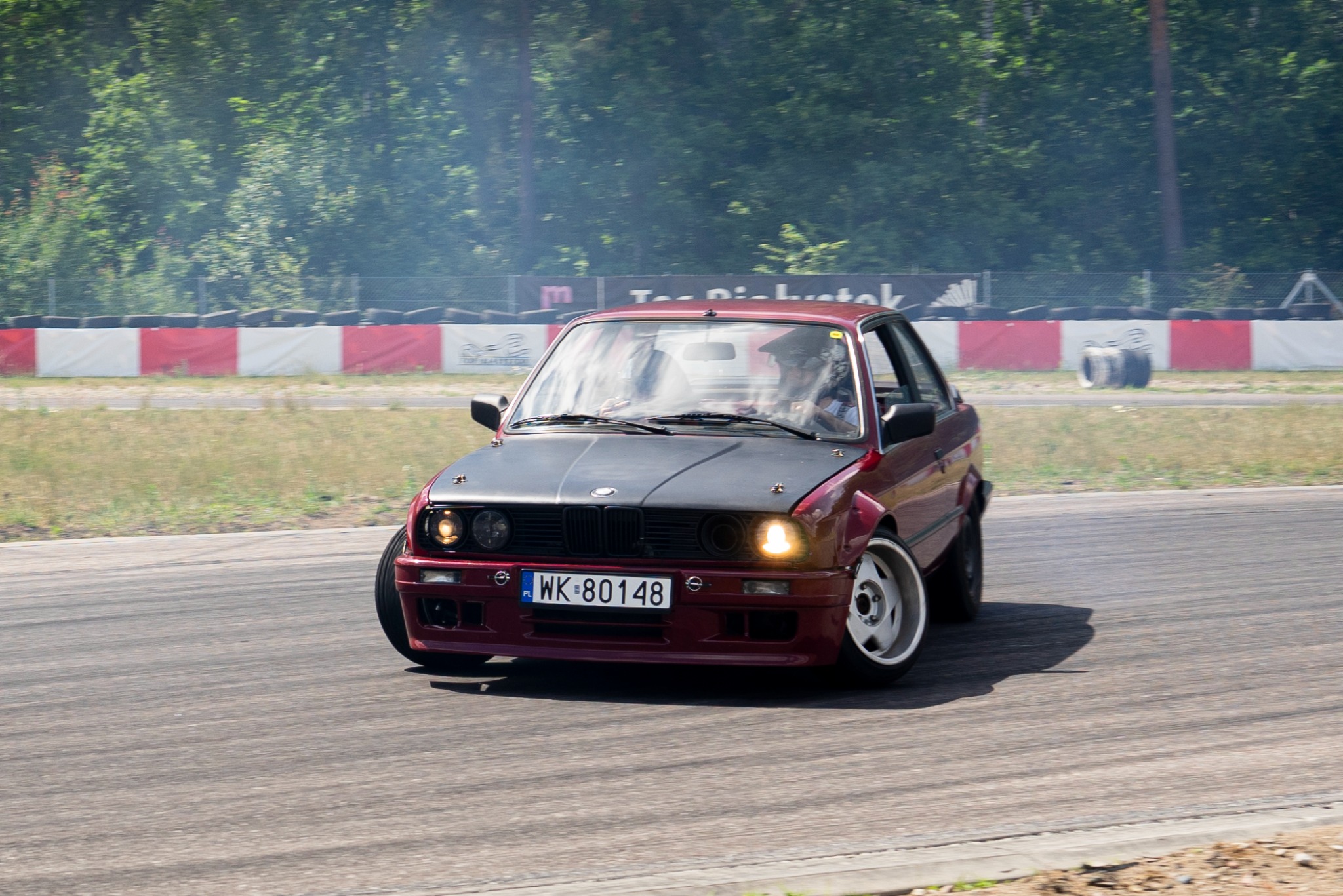 Czerwone BMW E30 z czarną maską, podczas driftu na torze. Widoczne opony z białym dymem, tablica rejestracyjna WK 80148, drzewa i opony w tle.