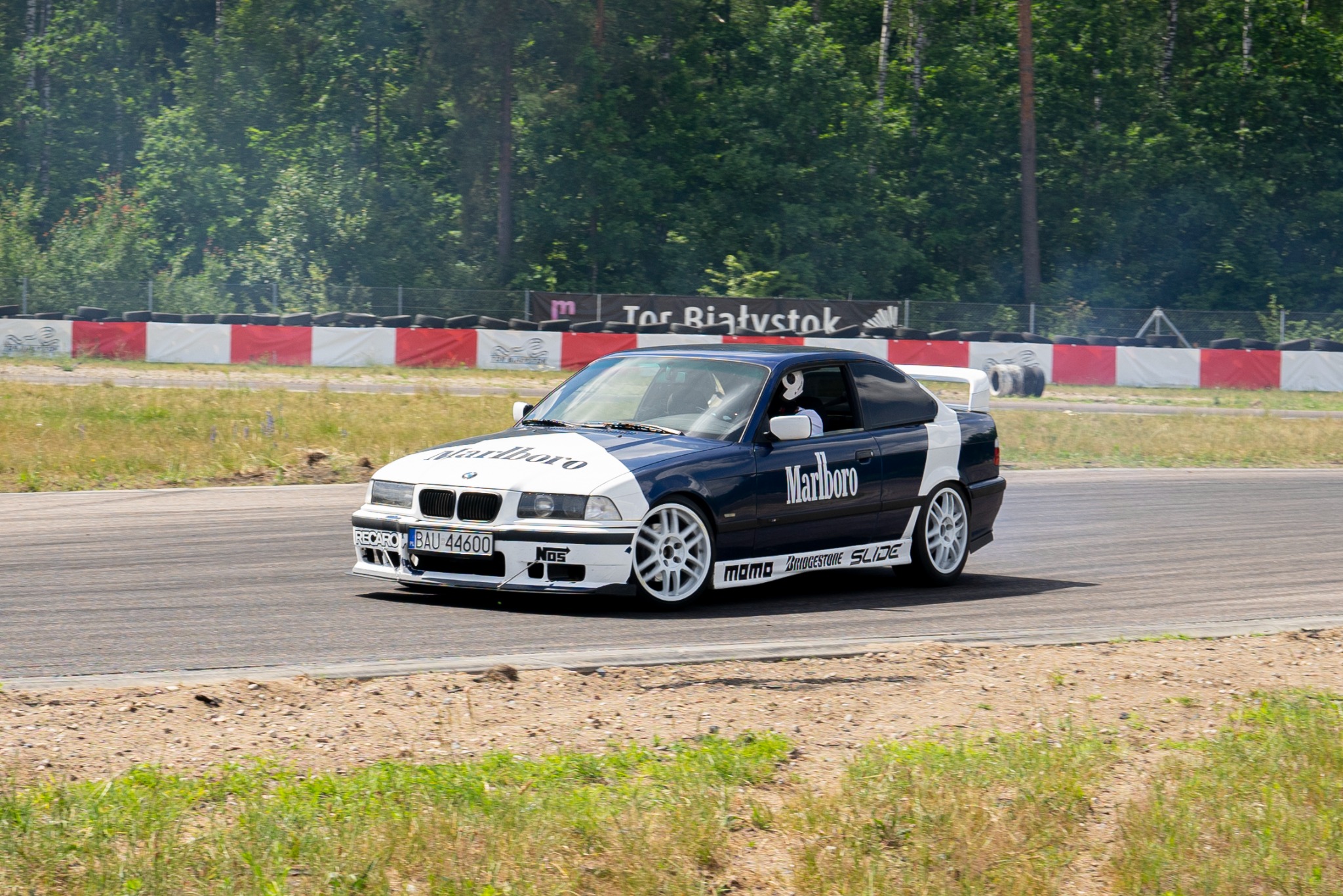 BMW E36 z oklejeniem Marlboro driftuje na torze Tor Białystok, w tle opony i zieleń. Biało-niebieska karoseria, białe felgi, widoczny kierowca w kasku.