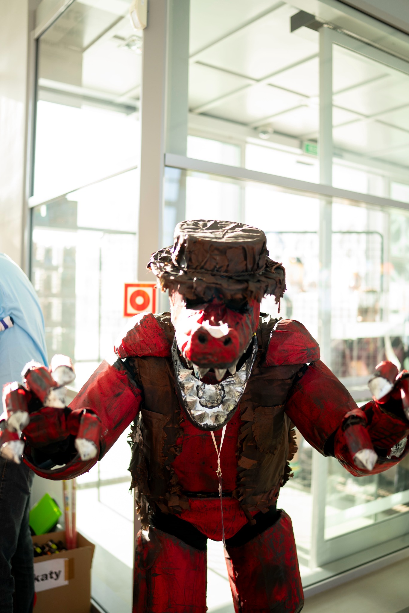 Osoba w ręcznie robionym, czerwono-brązowym kostiumie potwora z kapeluszem, pozująca na tle przeszklonego budynku. Kostium ma srebrne akcenty i ostre elementy.