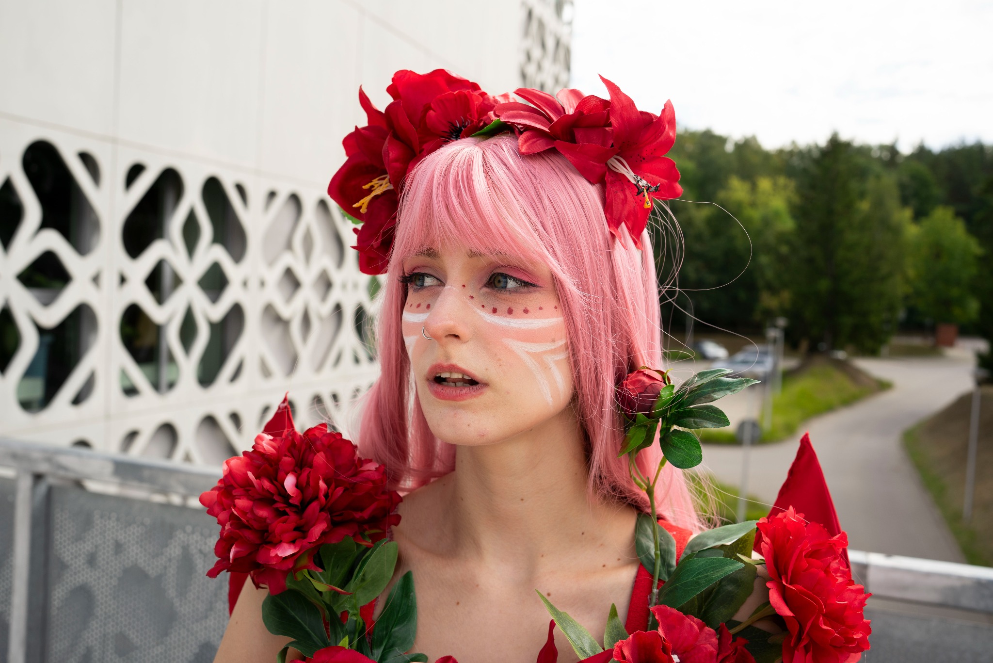 Portret kobiety z różowymi włosami, wiankiem z czerwonych kwiatów i białym makijażem na twarzy, na tle nowoczesnego budynku i zieleni.