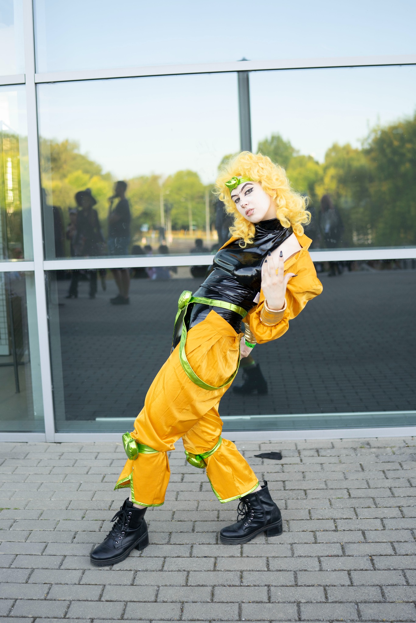 Osoba w kostiumie Dio Brando z JoJo's Bizarre Adventure, pozująca przed szklanym budynkiem. Żółty strój, czarne buty i blond peruka, widoczne sylwetki w tle.