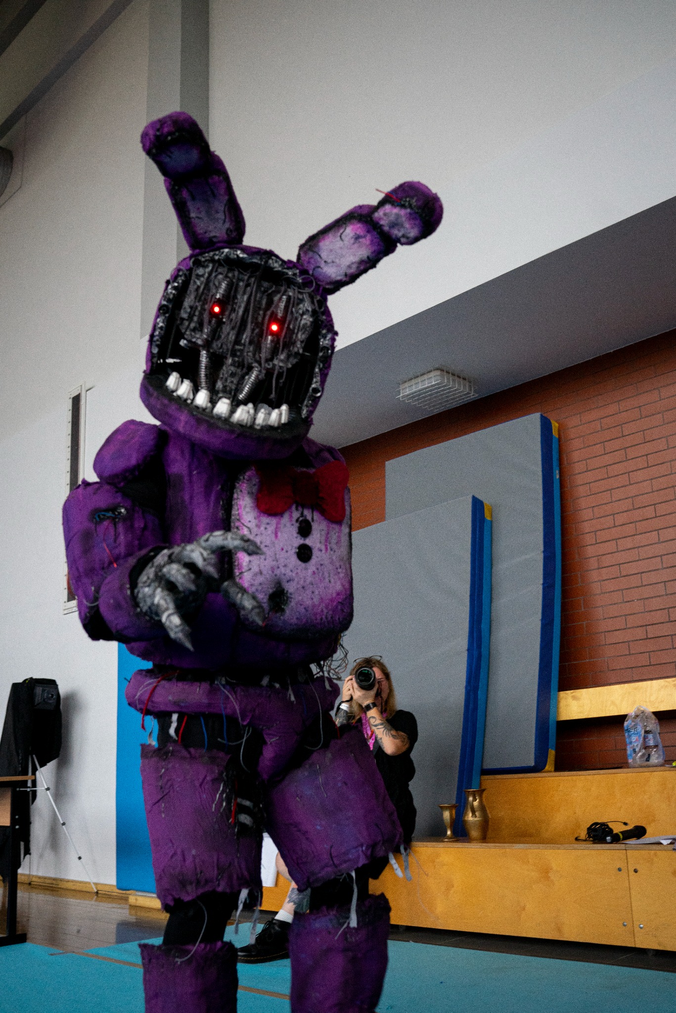 Fotograf uwiecznia postać w fioletowym kostiumie robota z czerwonymi oczami na tle sali gimnastycznej. Nietypowy cosplay i sesja zdjęciowa w sportowym otoczeniu.