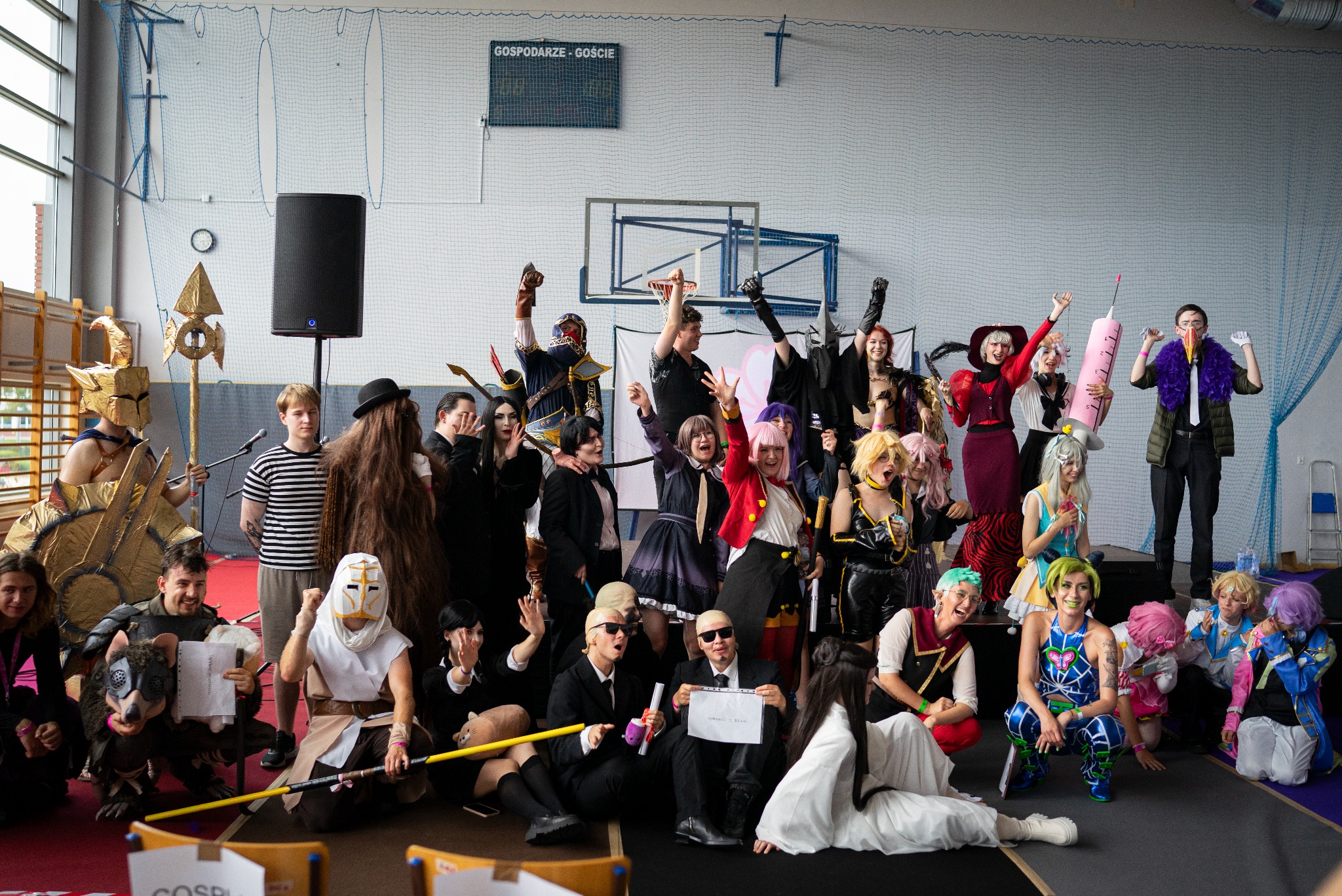 Grupowe zdjęcie uczestników konwentu cosplay w hali sportowej. Kostiumy postaci z gier i anime, różnorodne rekwizyty. W tle tablica wyników i siatka zabezpieczająca.