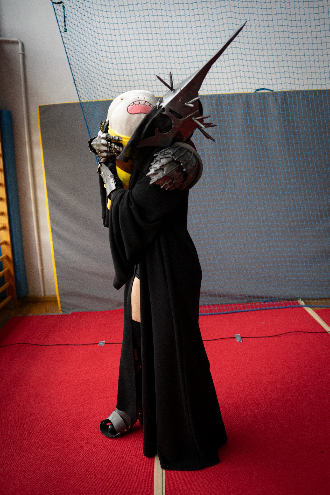 Osoba w czarnej szacie i hełmie z kolcami, trzymająca pluszową zabawkę, stoi na czerwonym dywanie w hali sportowej. Nietypowy kostium.
