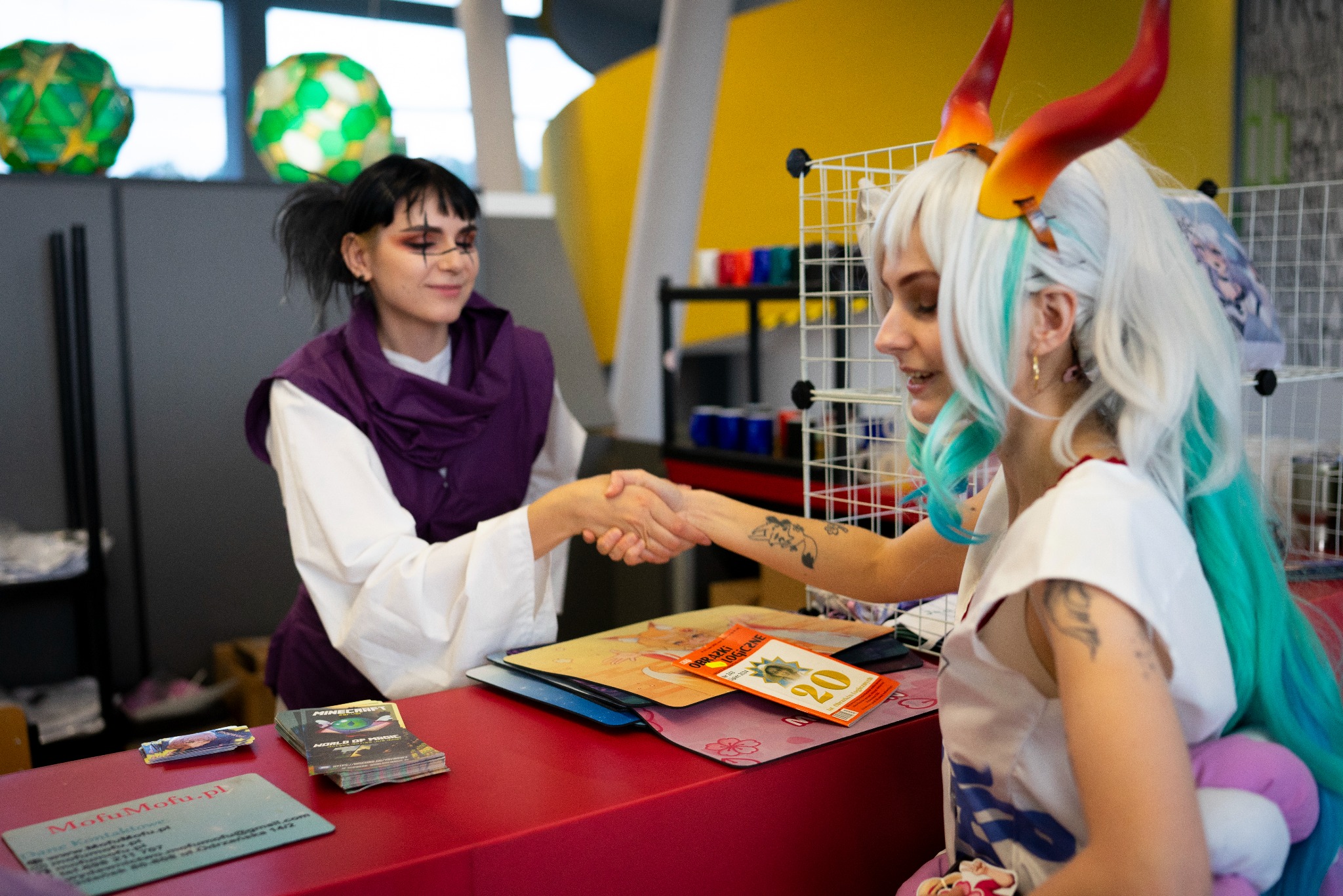 Dwie osoby w kostiumach wymieniają uścisk dłoni za czerwonym stołem z akcesoriami. Jedna osoba ma rogi i biało-turkusowe włosy, druga ma ciemne włosy i fioletowy szal.