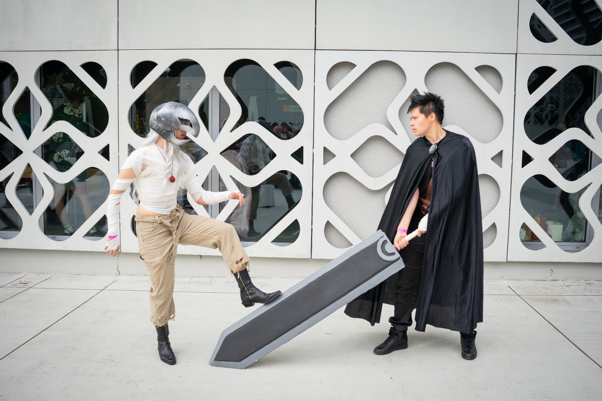 Dwie osoby w kostiumach cosplay, jedna z mieczem, pozują na tle nowoczesnej, białej fasady budynku z geometrycznymi wzorami.