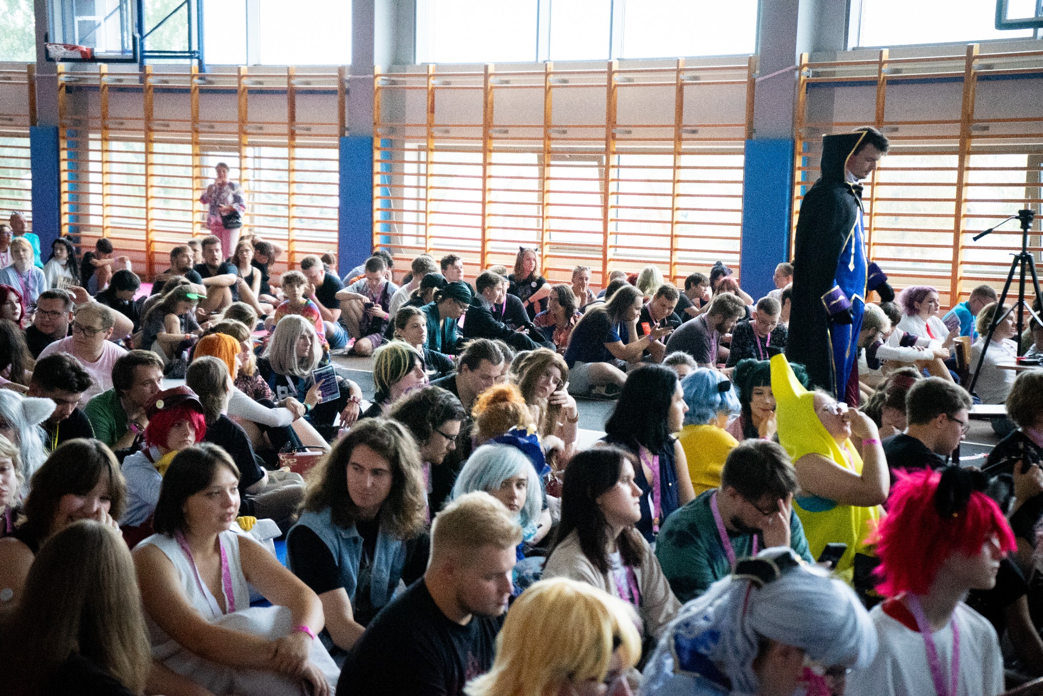 Spora grupa osób siedzi na podłodze w hali sportowej, prawdopodobnie podczas wydarzenia cosplay lub konwentu. W tle widoczne okna i drabinki gimnastyczne.