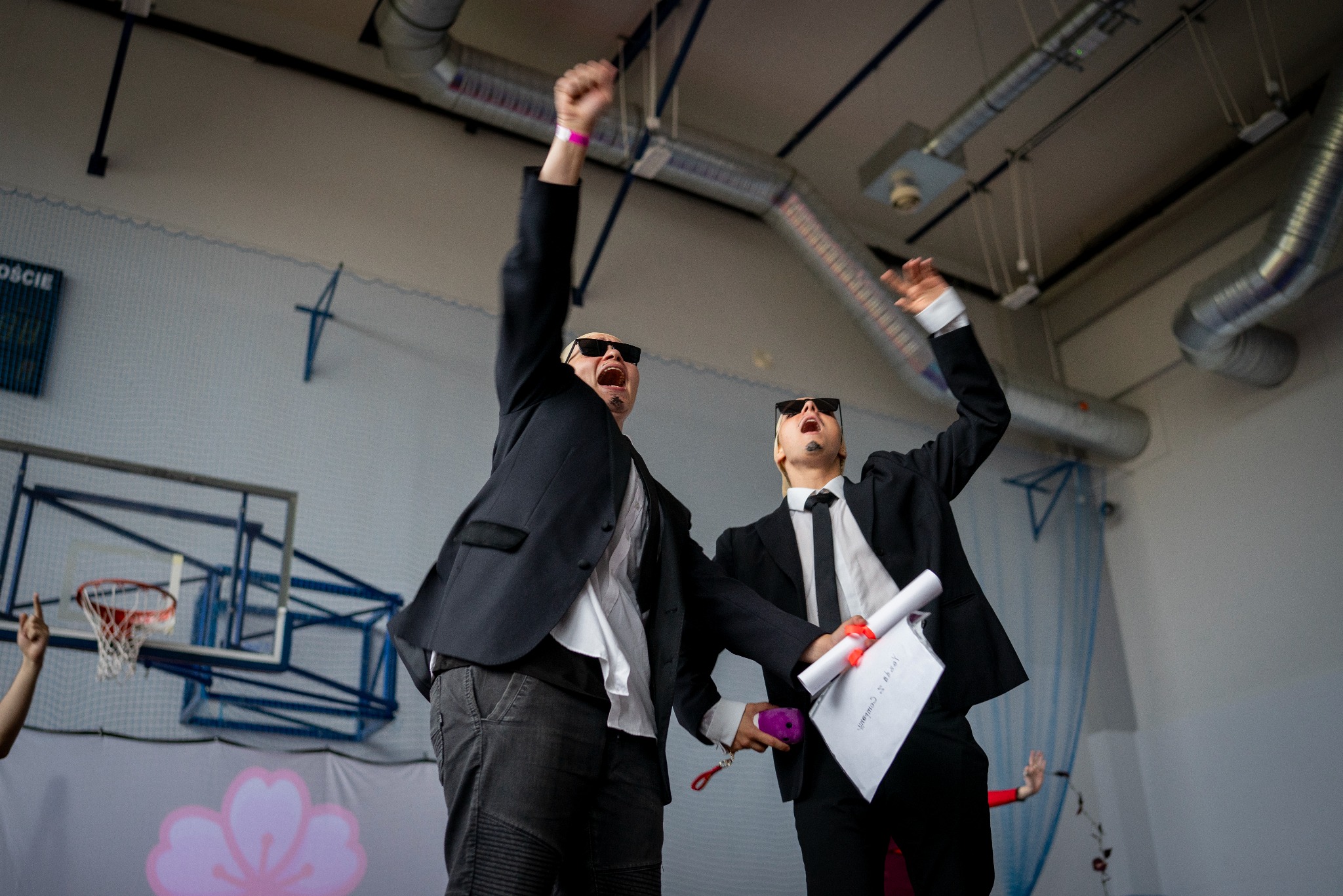 Ekscytacja w Białymstoku: Dwóch elegancko ubranych mężczyzn w okularach przeciwsłonecznych celebruje sukces, unosząc ręce w górę na tle sali gimnastycznej.