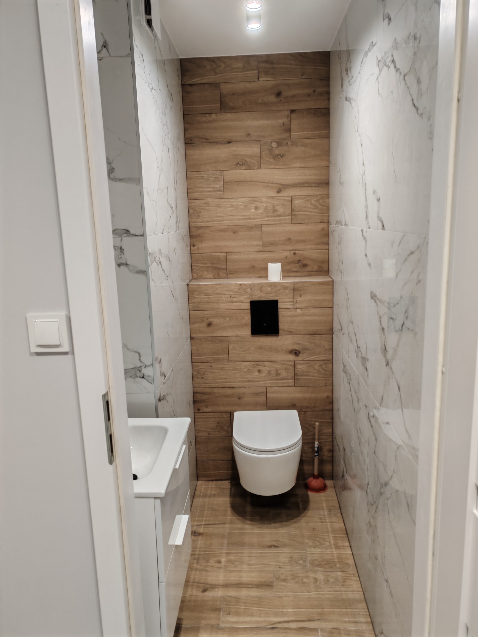 Wąska toaleta po remoncie: drewnopodobne i marmurowe płytki, podwieszana miska WC, umywalka z szafką. Widok zza uchylonych drzwi.
