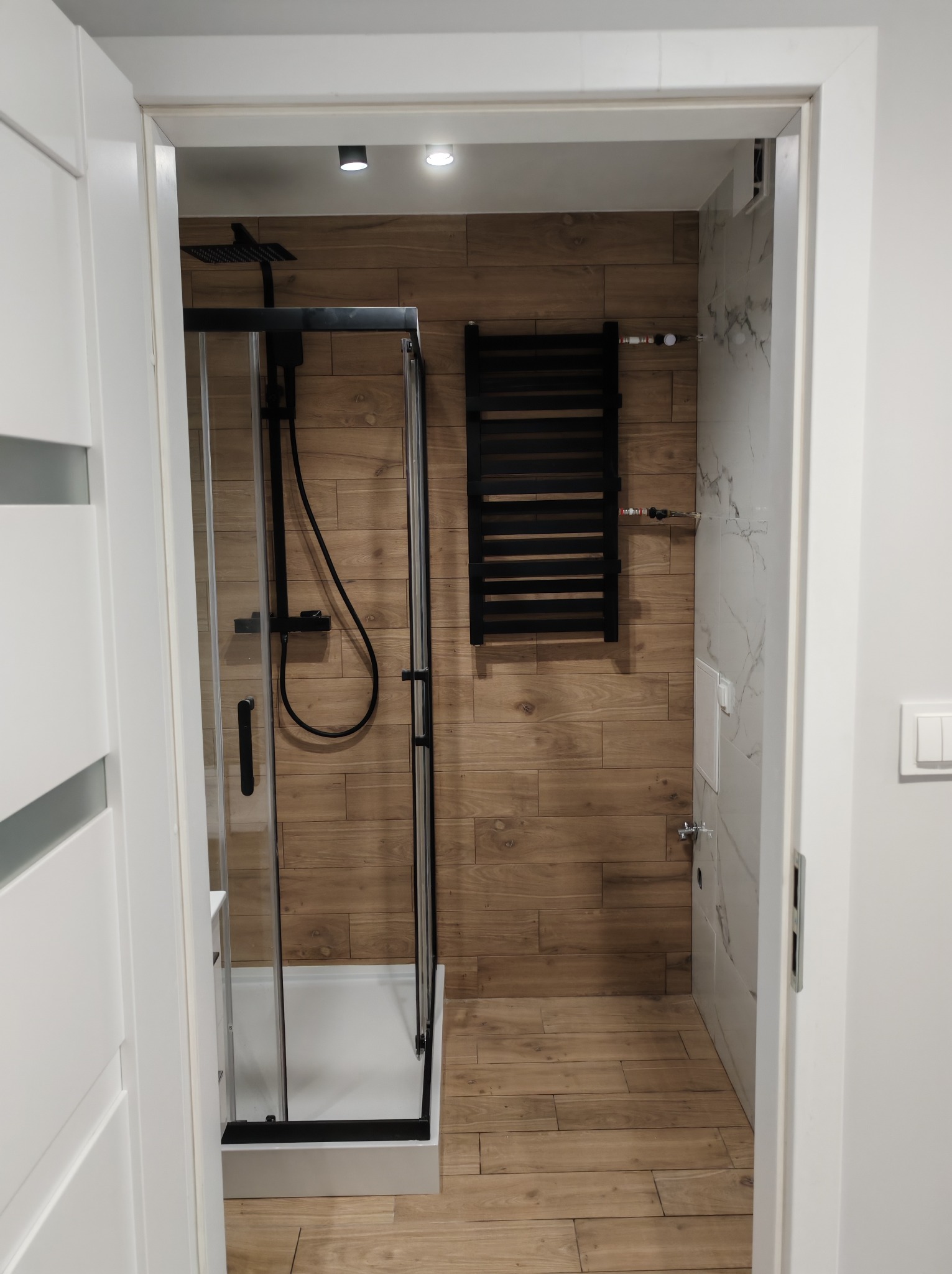 Nowoczesna łazienka po remoncie: kabina prysznicowa z czarnym wykończeniem, grzejnik drabinkowy i płytki imitujące drewno. Widok zza otwartych, białych drzwi.