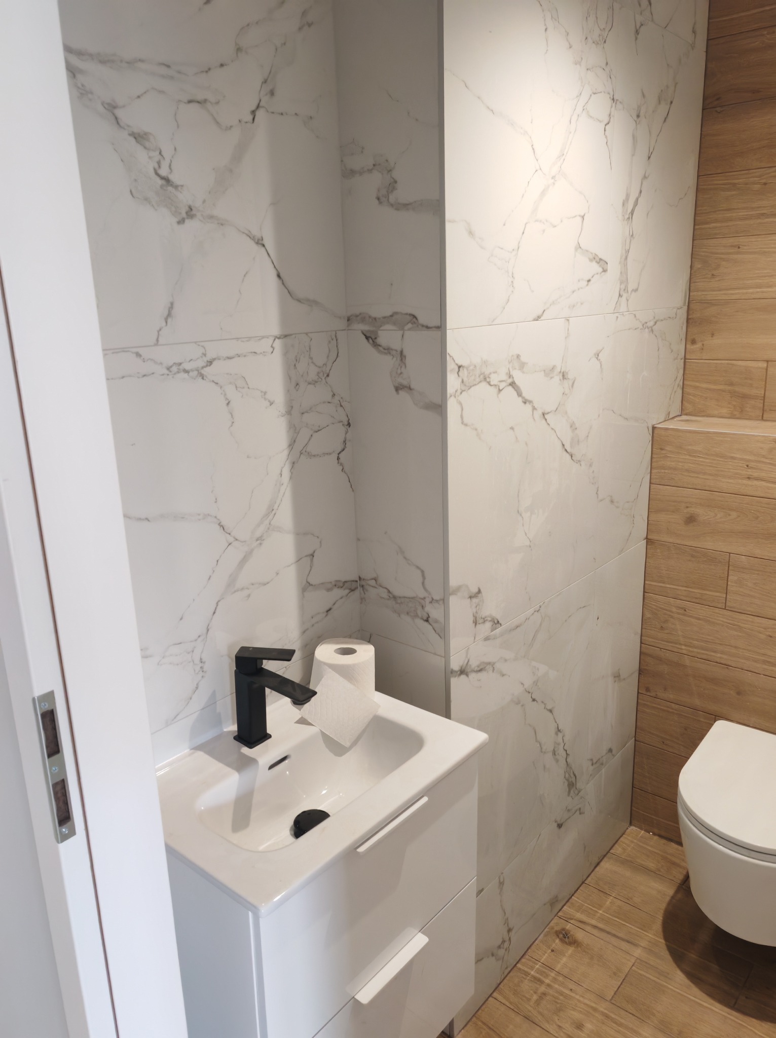 Nowoczesna łazienka w Kaliszu: biała umywalka z czarną armaturą, marmurowe płytki ścienne i drewnopodobna podłoga, tworzą eleganckie wykończenie wnętrza.