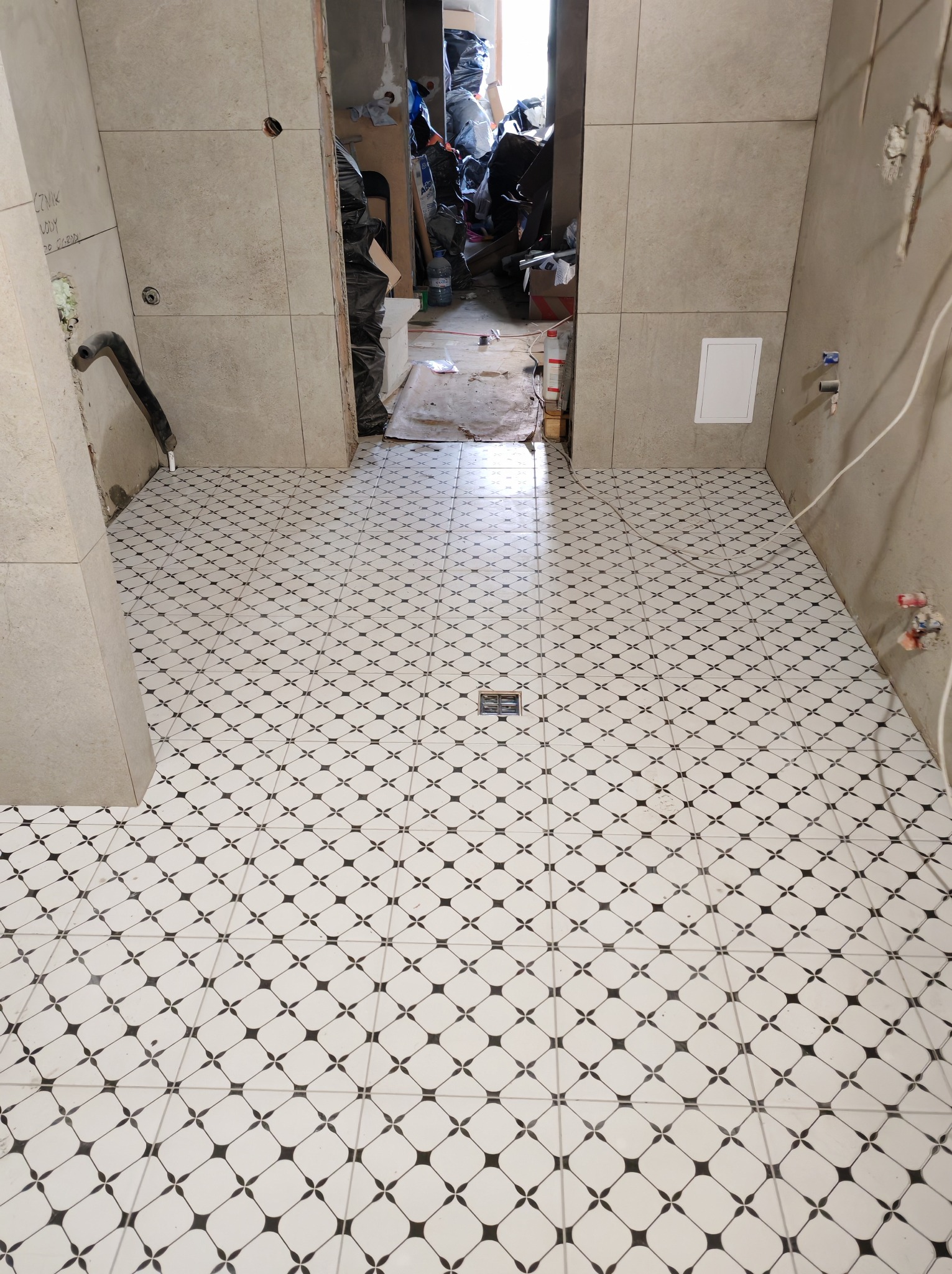 Ułożona podłoga z płytek w biało-czarny wzór w łazience w trakcie remontu. Widoczny odpływ liniowy i fragmenty ścian z beżowych płytek.