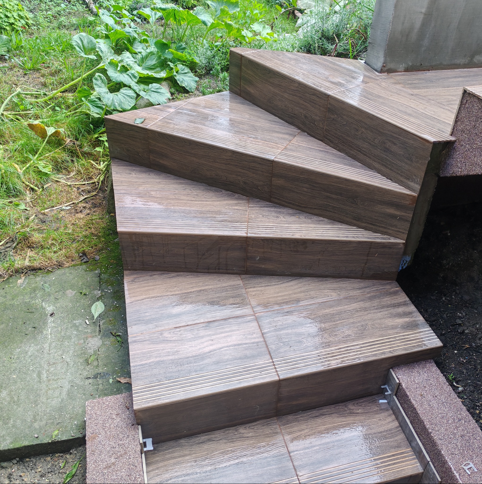 Schody zewnętrzne wyłożone brązowymi płytkami imitującymi drewno, z widocznymi elementami montażowymi i zielenią w tle. Detal wykończenia schodów.